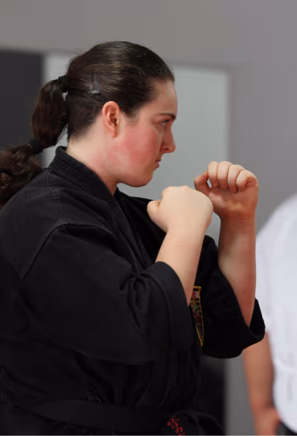 An adult student in a black karate gi stands ready and on guard as she practices techniques with her sensei during karate class.