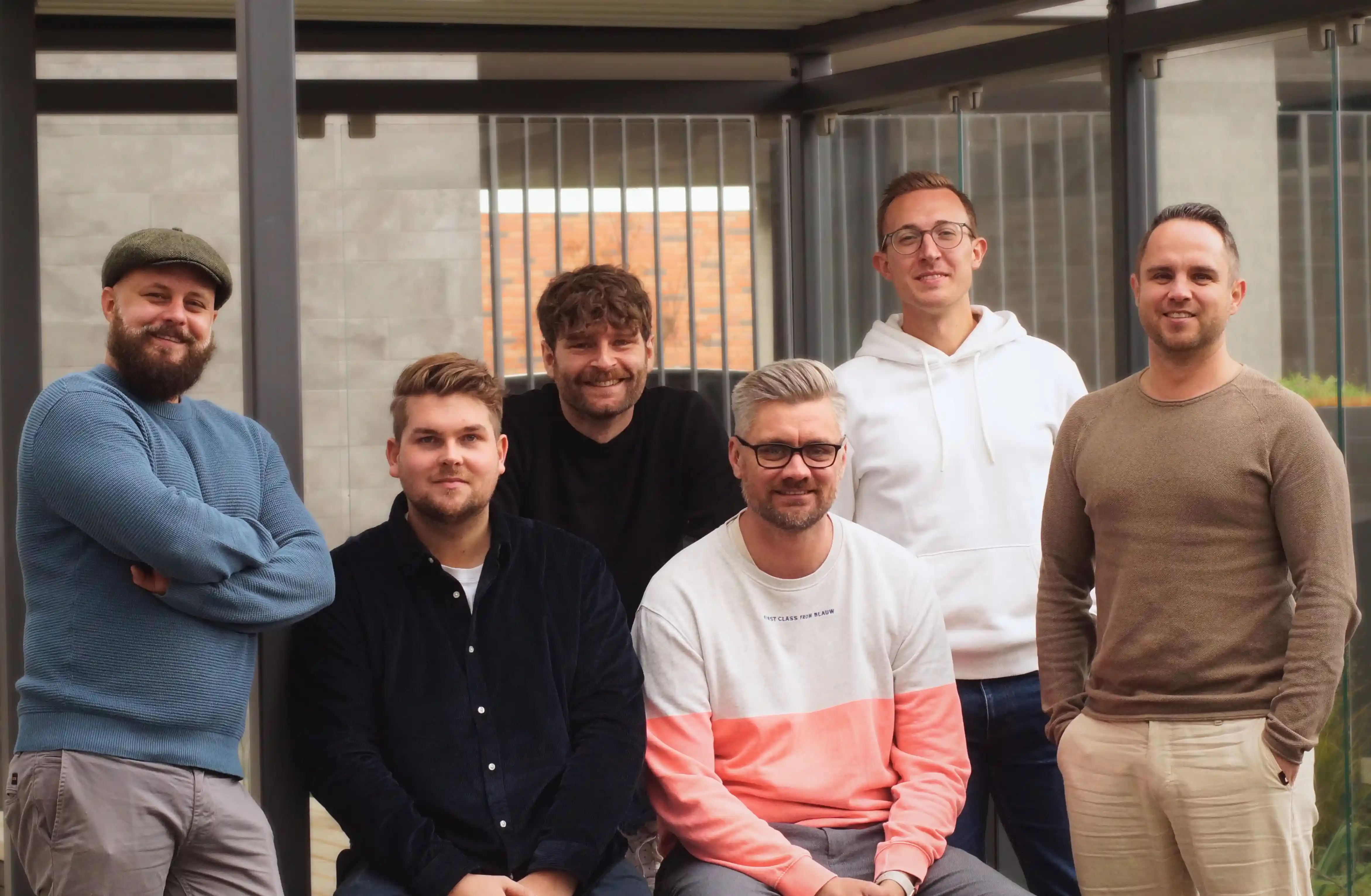 Vier Männer sitzen entspannt auf einem dunklen Sofa in einem modern eingerichteten Büro mit LBX-Logo an der Wand. Sie lächeln in die Kamera, im Vordergrund stehen zwei runde Couchtische
