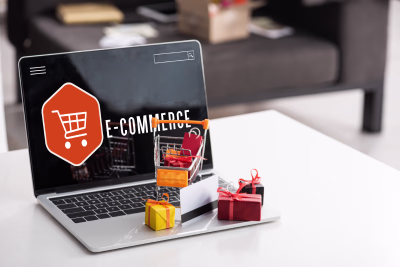 Laptop displaying "E-Commerce" with a shopping cart icon. Miniature gifts and cart on keyboard, symbolizing online shopping's convenience. Bright, inviting scene.