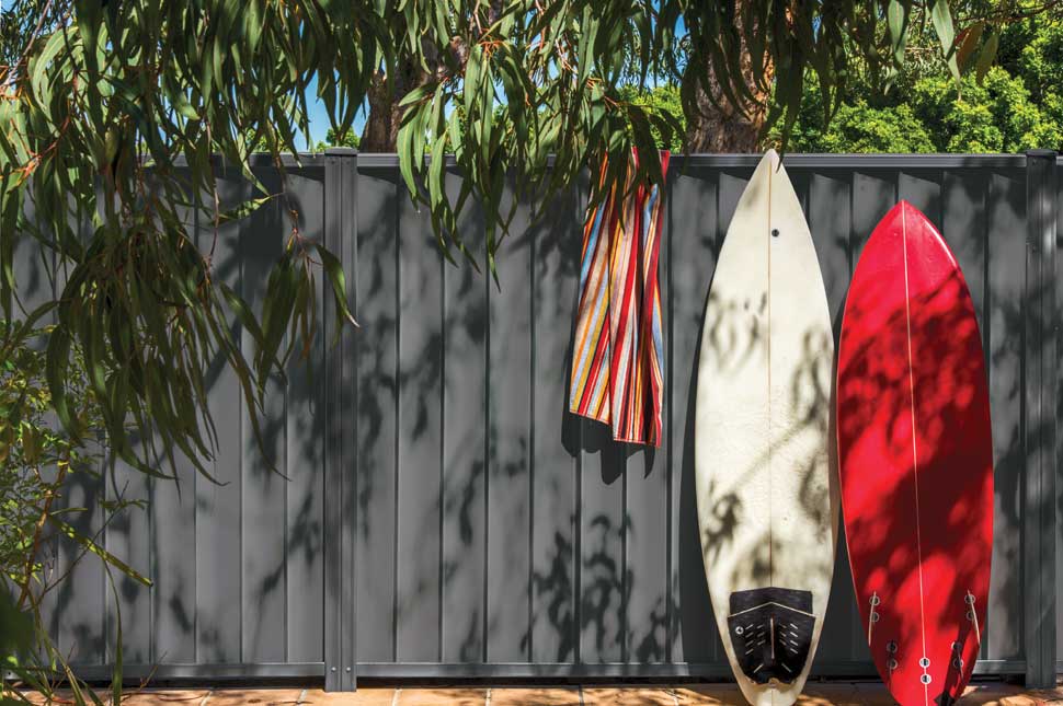 Dark gray Colorbond steel fence with vertical panels, featuring two surfboards and a colorful striped towel hanging in front