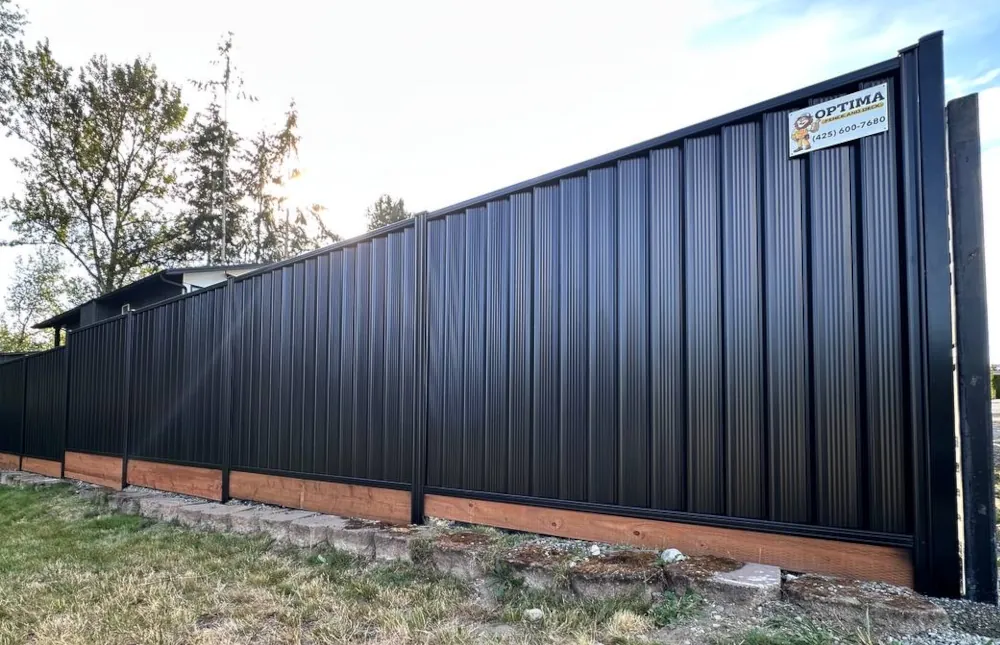 Black corrugated metal privacy fence with wood base trim along a grassy yard