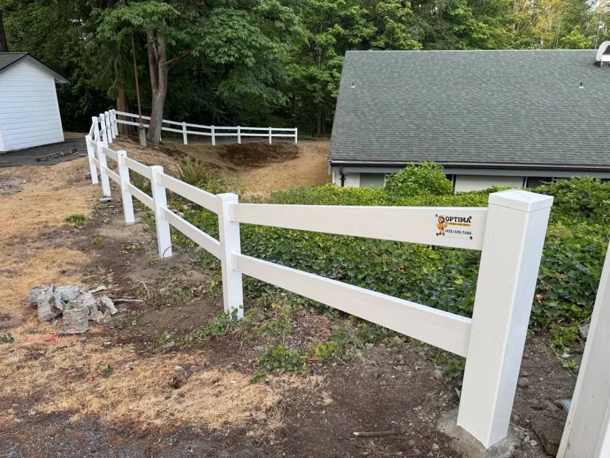 White vinyl ranch rail fence installed along a sloped property with dirt ground and trees by Optima Fence and Deck in the Bellevue area