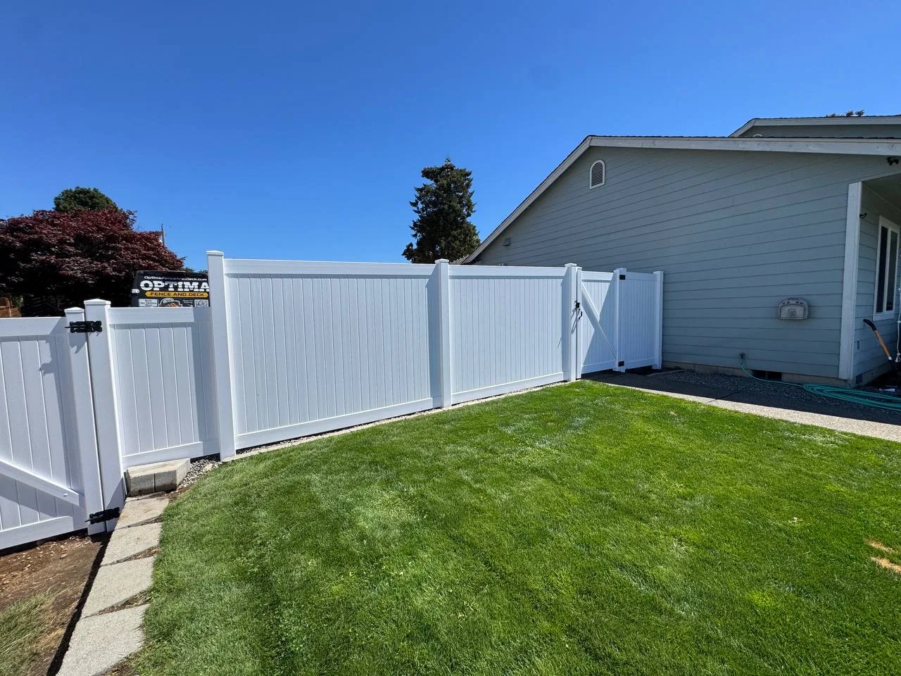 White vinyl privacy fence with gate section installed by Optima Fence and Deck at a blue-gray sided home with a green lawn in Bellevue