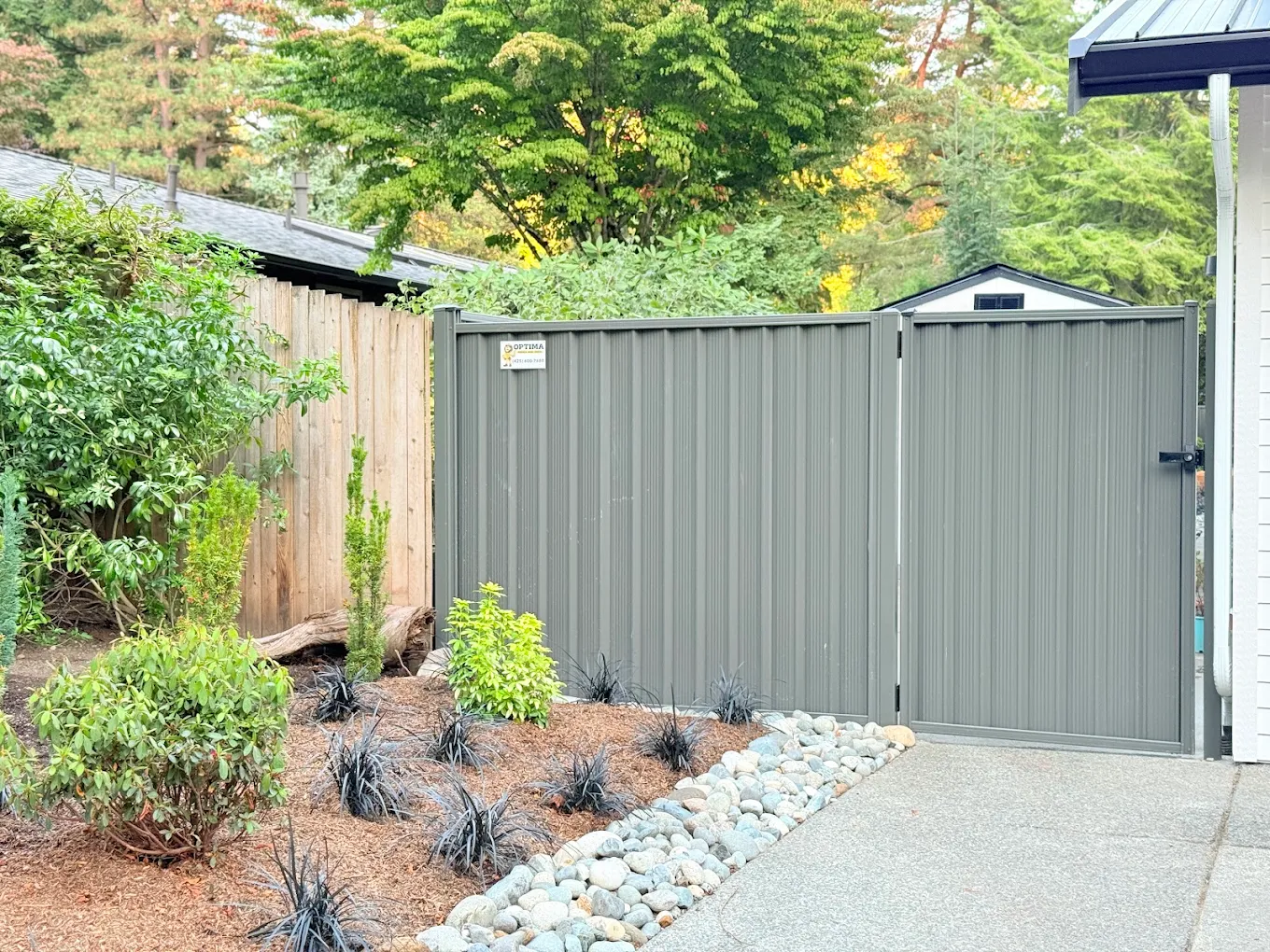 Gray vinyl privacy fence with walk-through gate in a professionally landscaped yard featuring decorative rocks, bark mulch, and ornamental shrubs