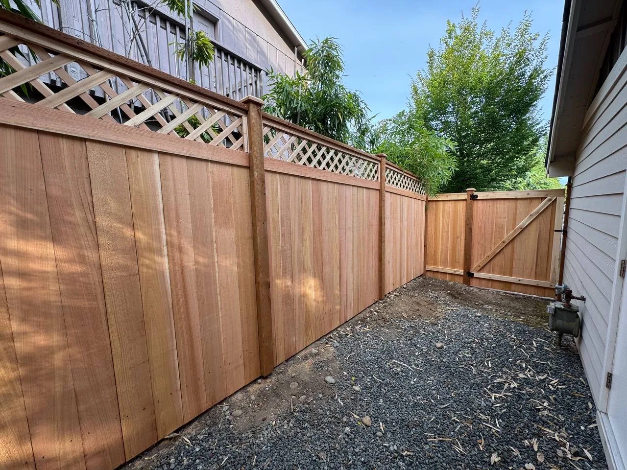 Cedar-toned faux wood vinyl fence with decorative lattice top and vertical board design running along a gravel side yard pathway beside a residential home in Bellevue