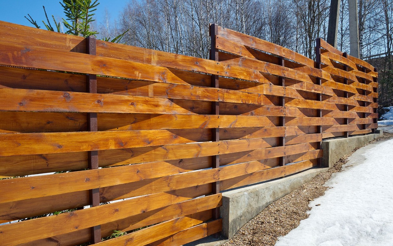 Modern horizontal wood fence with warm stained boards installed along a concrete base beside a snowy yard