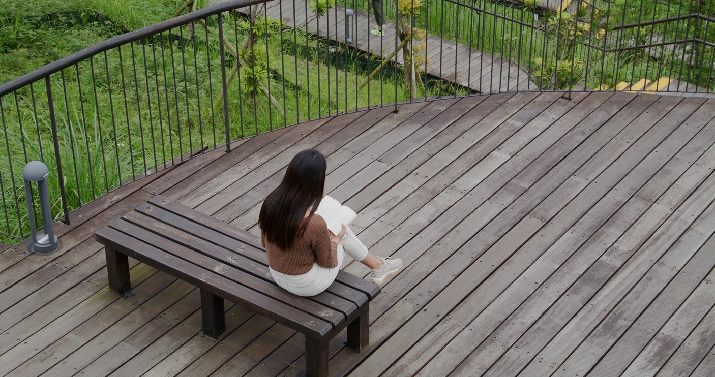 Woman sitting on a bench reading a book on a wood deck with metal railing