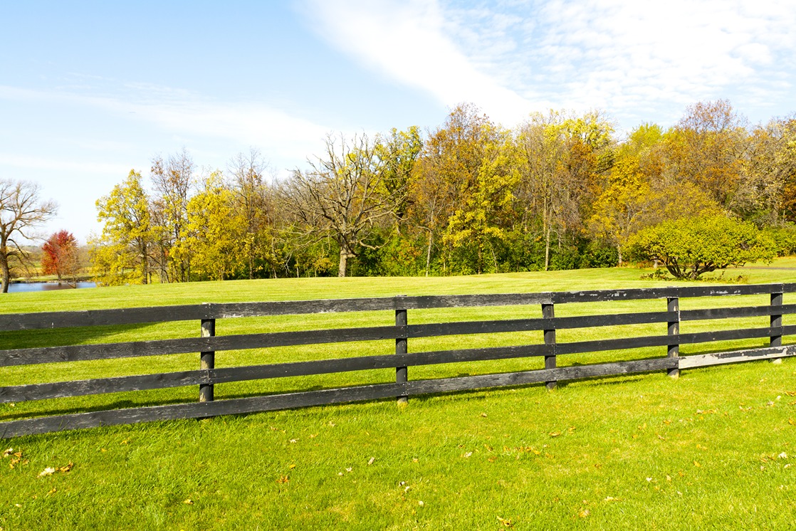 Black four rail wood fence bordering an open pasture with autumn trees