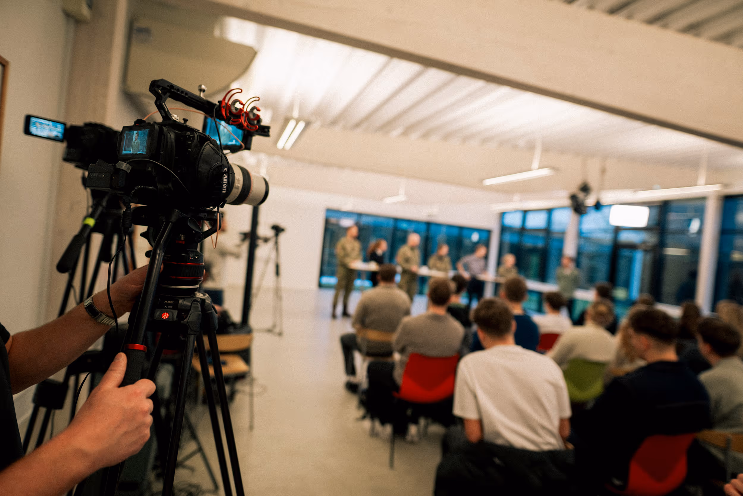 Video camera recording a panel discussion with several people seated and speaking in a modern room.