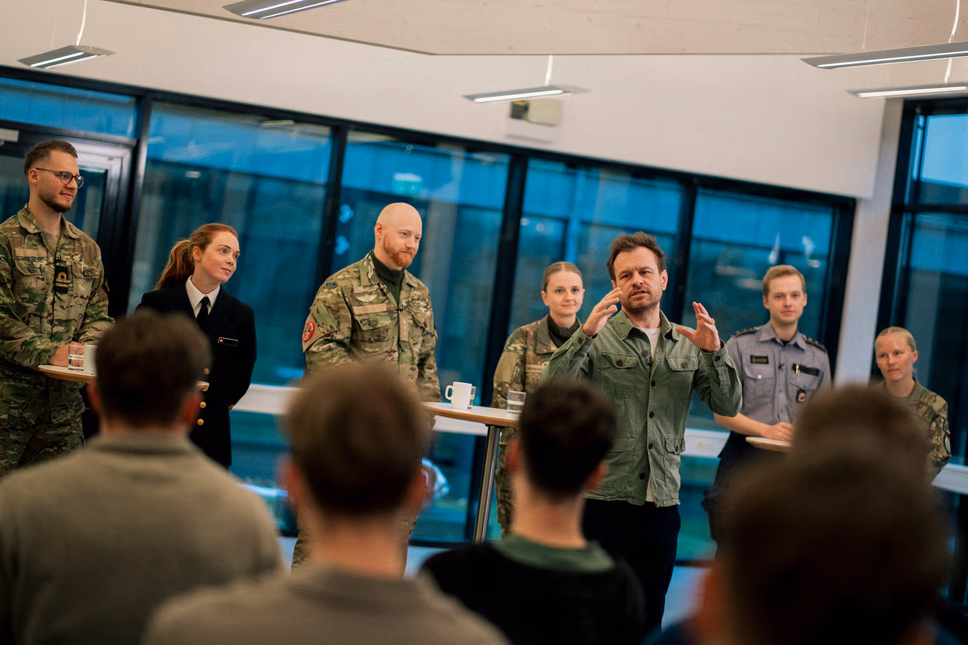 A group of military personnel and a man in a green jacket speaking to an audience in a modern indoor setting.