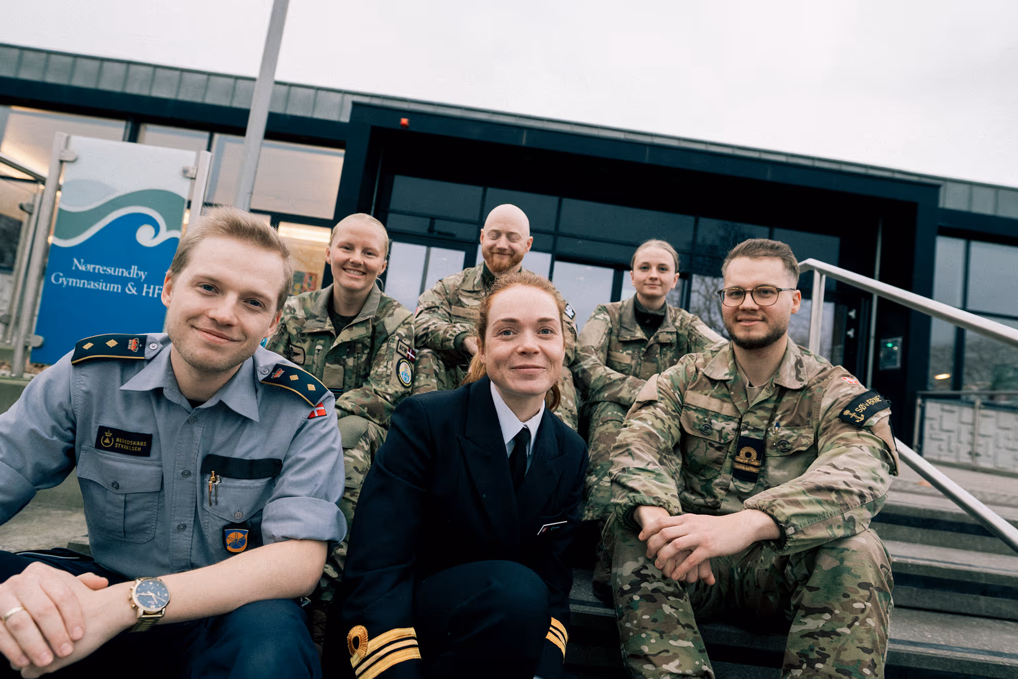 Group of six smiling people in military and emergency service uniforms sitting on steps outside a building with a sign reading Nørresundby Gymnasium & HF.