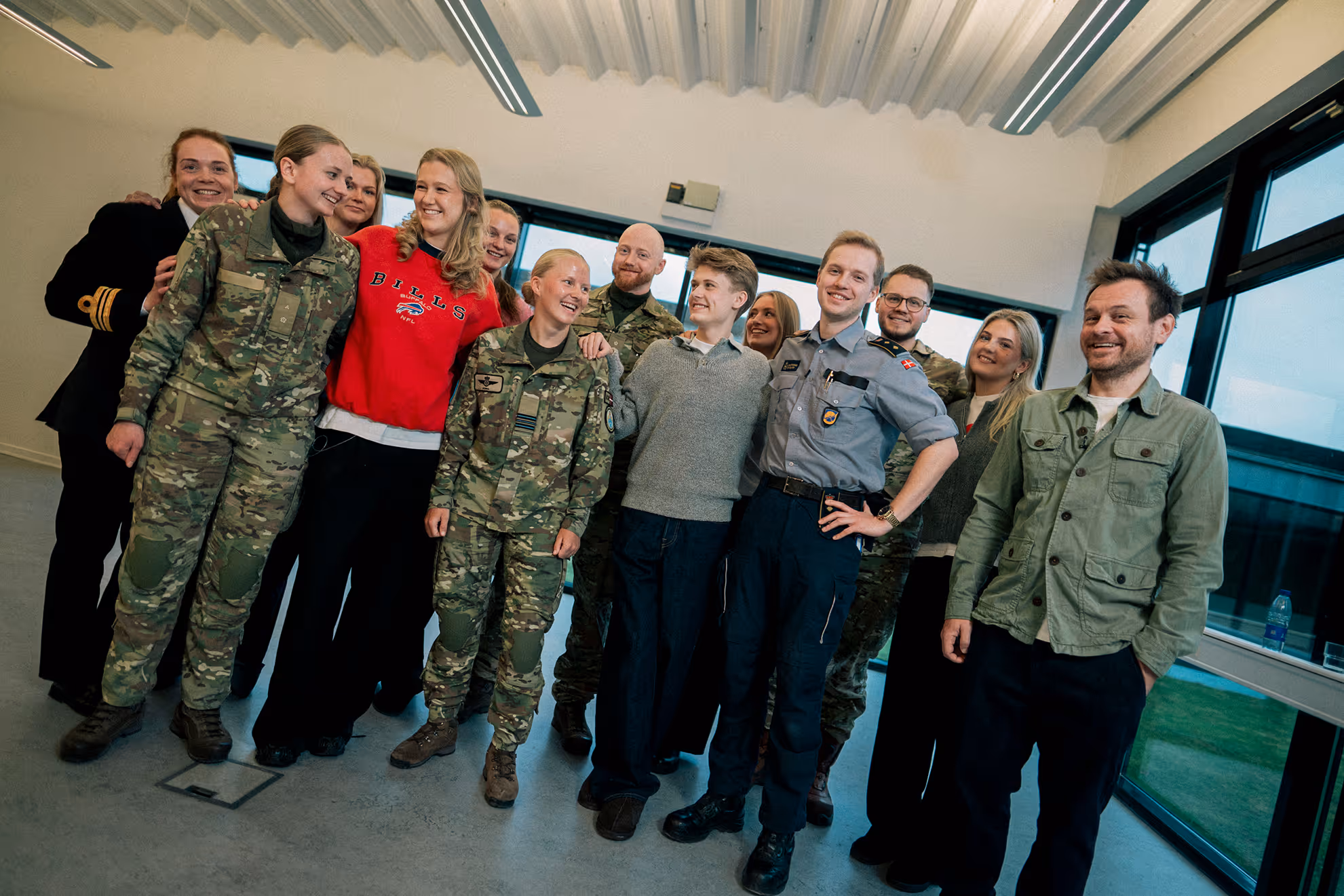 Group of smiling people, including individuals in military uniforms and casual clothing, standing together inside a room with large windows.