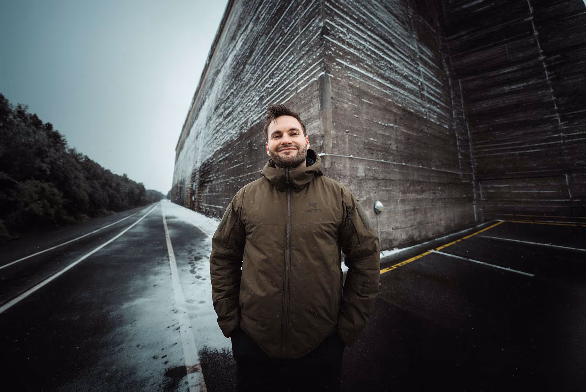Group of smiling people, including individuals in military uniforms and casual clothing, standing together inside a room with large windows.