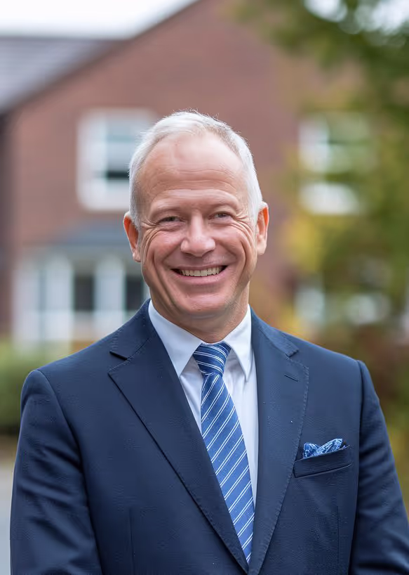 Bald man with a broad smile wearing a dark blue suit and striped tie, standing outdoors with a blurred residential building in the background, likely a professional portrait for business or social use