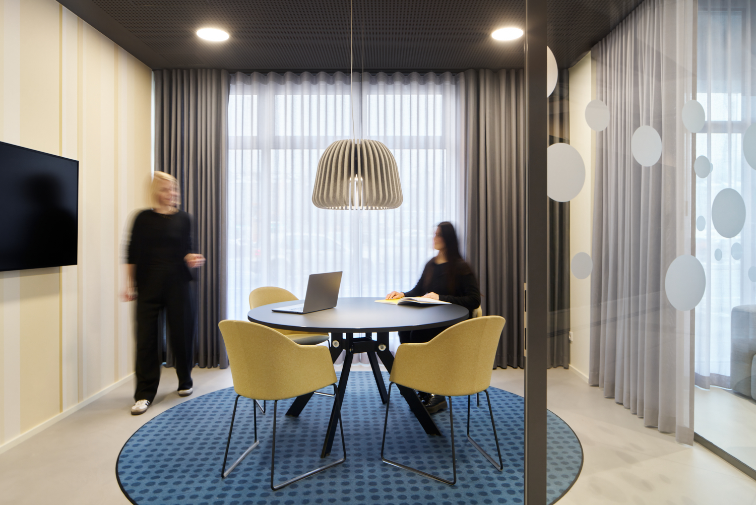 Moderner Besprechungsraum mit rundem Tisch, gelben Stühlen, Laptop und zwei verschwommenen Frauen, eine sitzt und liest, die andere steht.