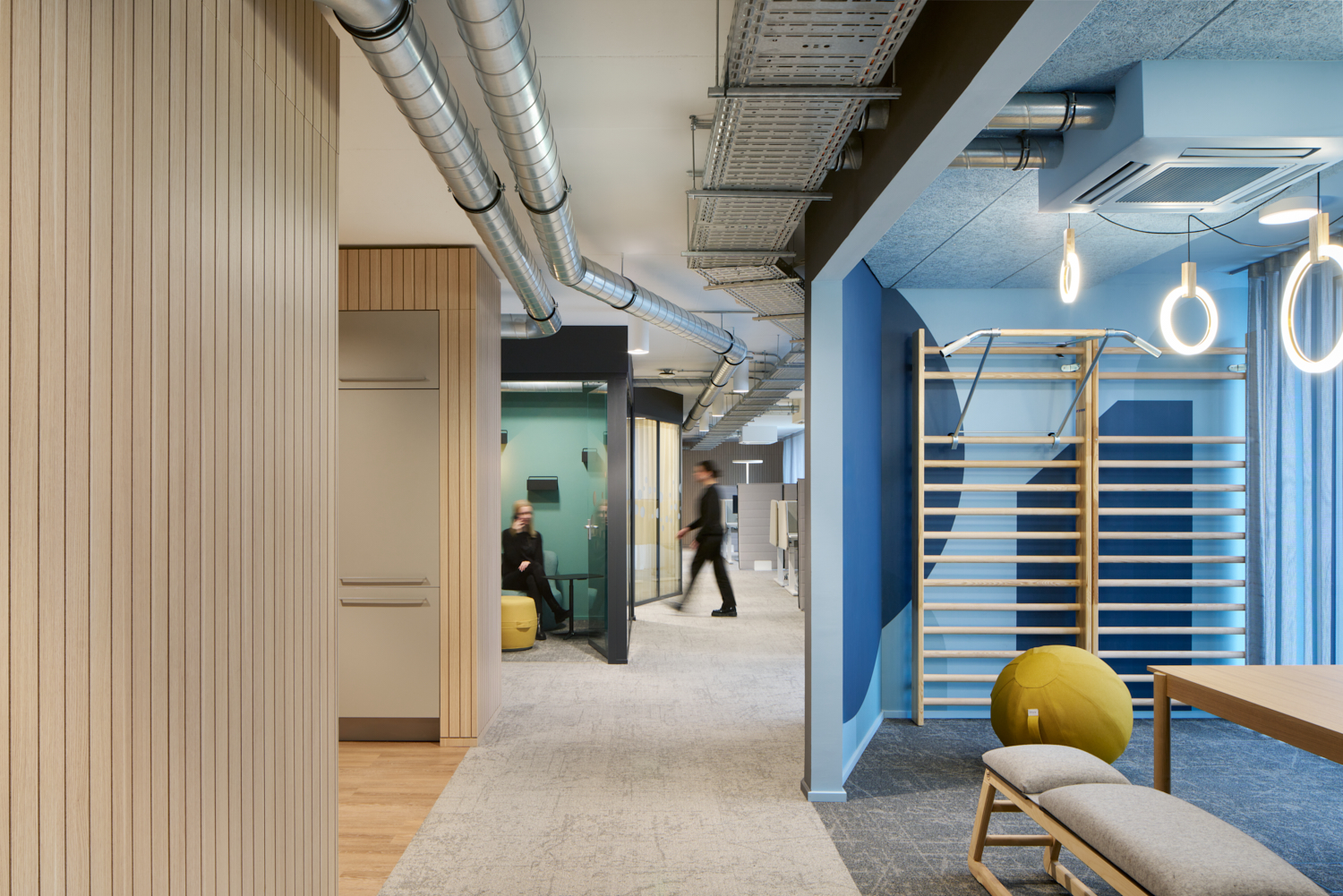 Modernes Büro mit offenen Arbeitsbereichen, Holzverkleidungen, sichtbaren Lüftungsrohren und blauen Wandelementen mit Fitnessgeräten.