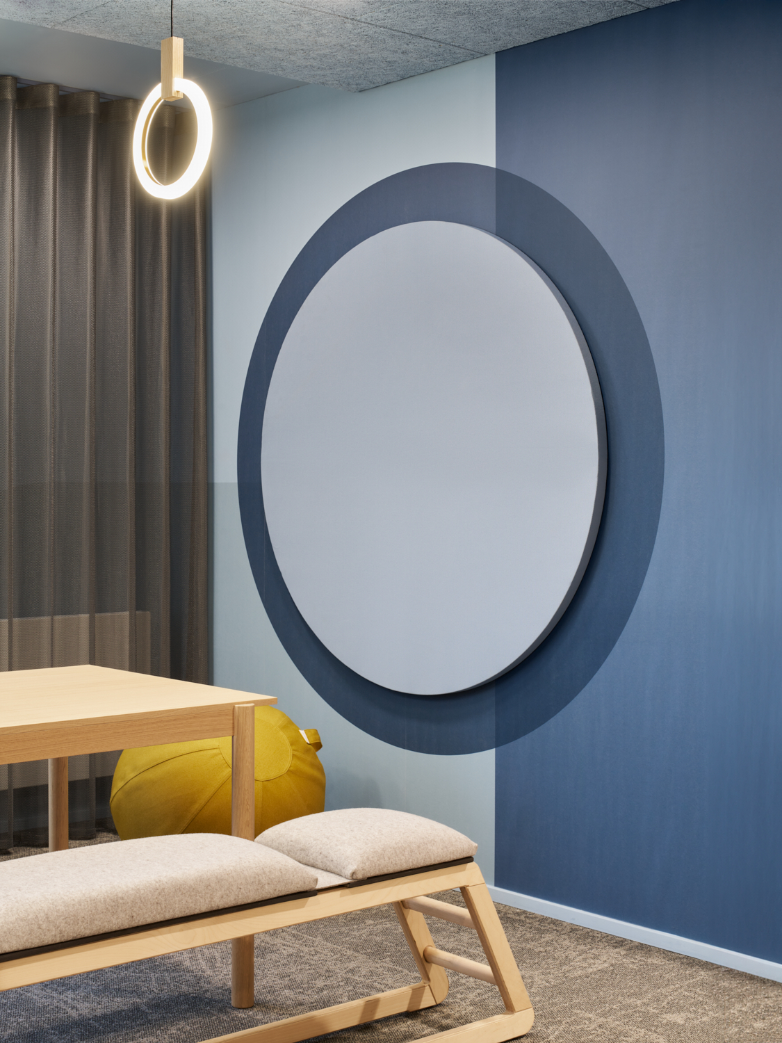 Moderner Innenraum mit holzfarbenem Tisch, gepolsterter Bank, gelbem Sitzsack, runder Spiegel an der zweifarbigen blauen Wand und ringförmiger Deckenleuchte.