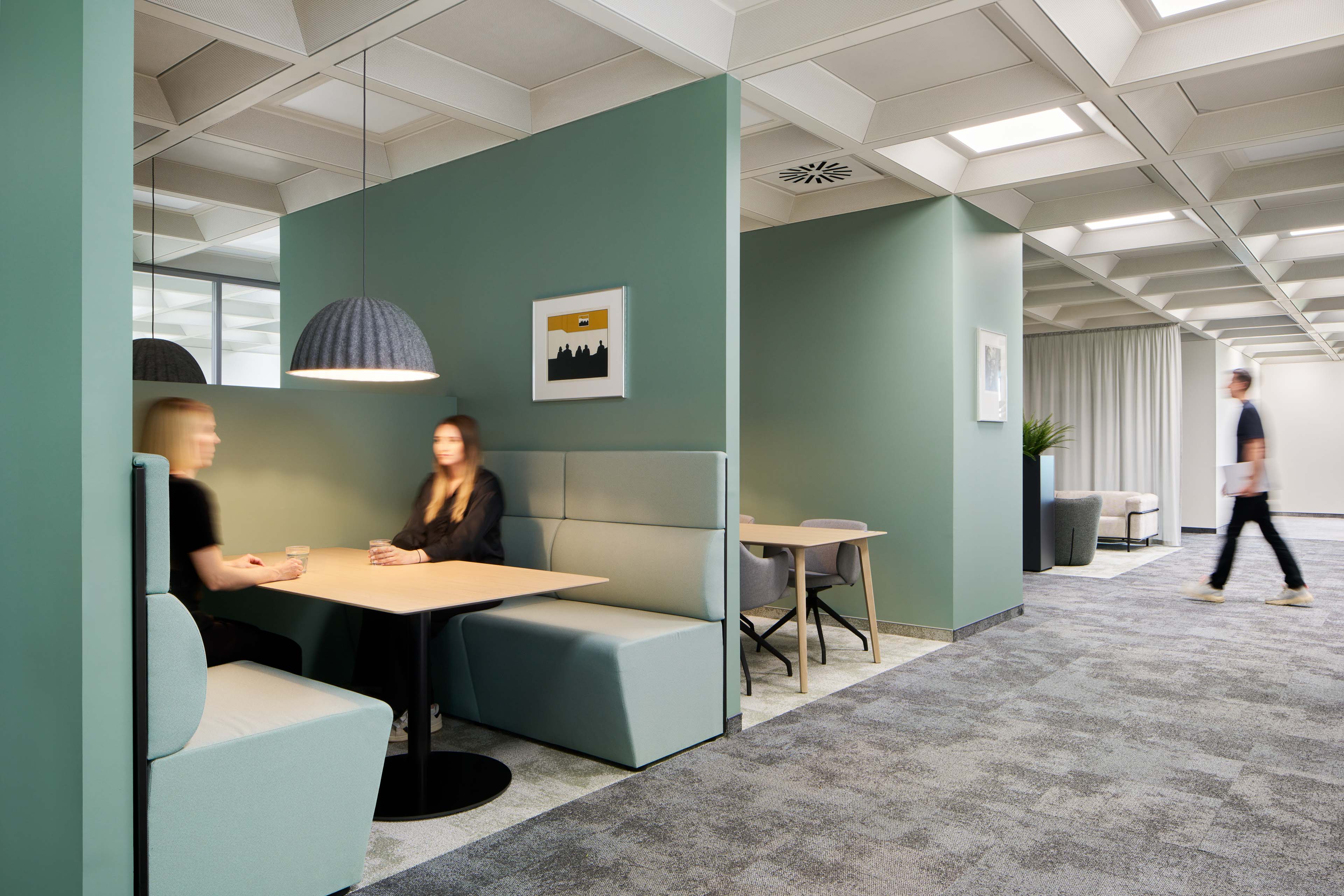 Moderner Bürobereich mit Sitzecke, in der zwei Frauen sich gegenüber an einem Tisch unter einer Hängeleuchte unterhalten, und eine Person, die mit Laptop vorbei geht.