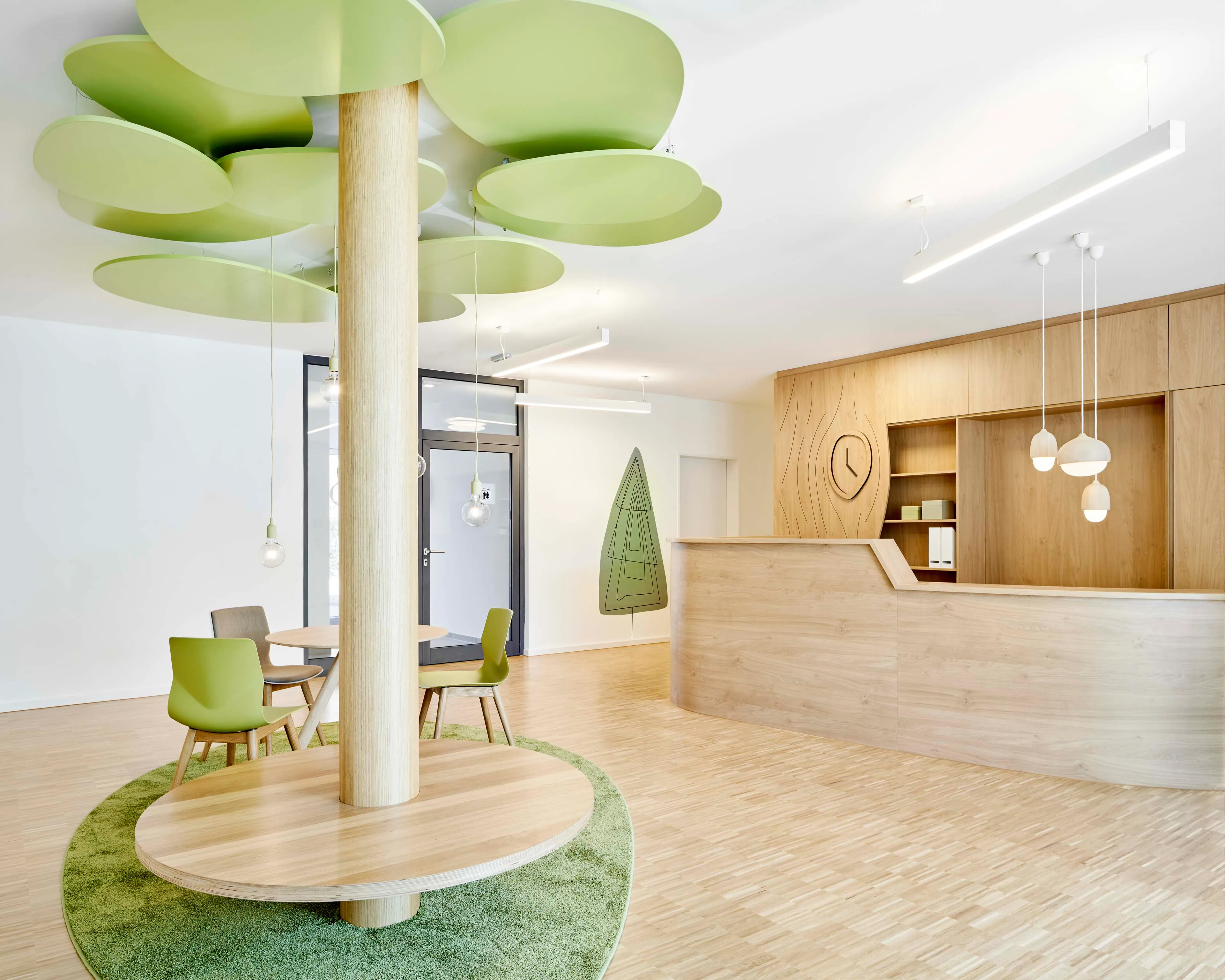 Hell eingerichteter Empfangsbereich mit grünem Teppich und Stühlen, hölzerner Theke und Baum-Design-Decke.