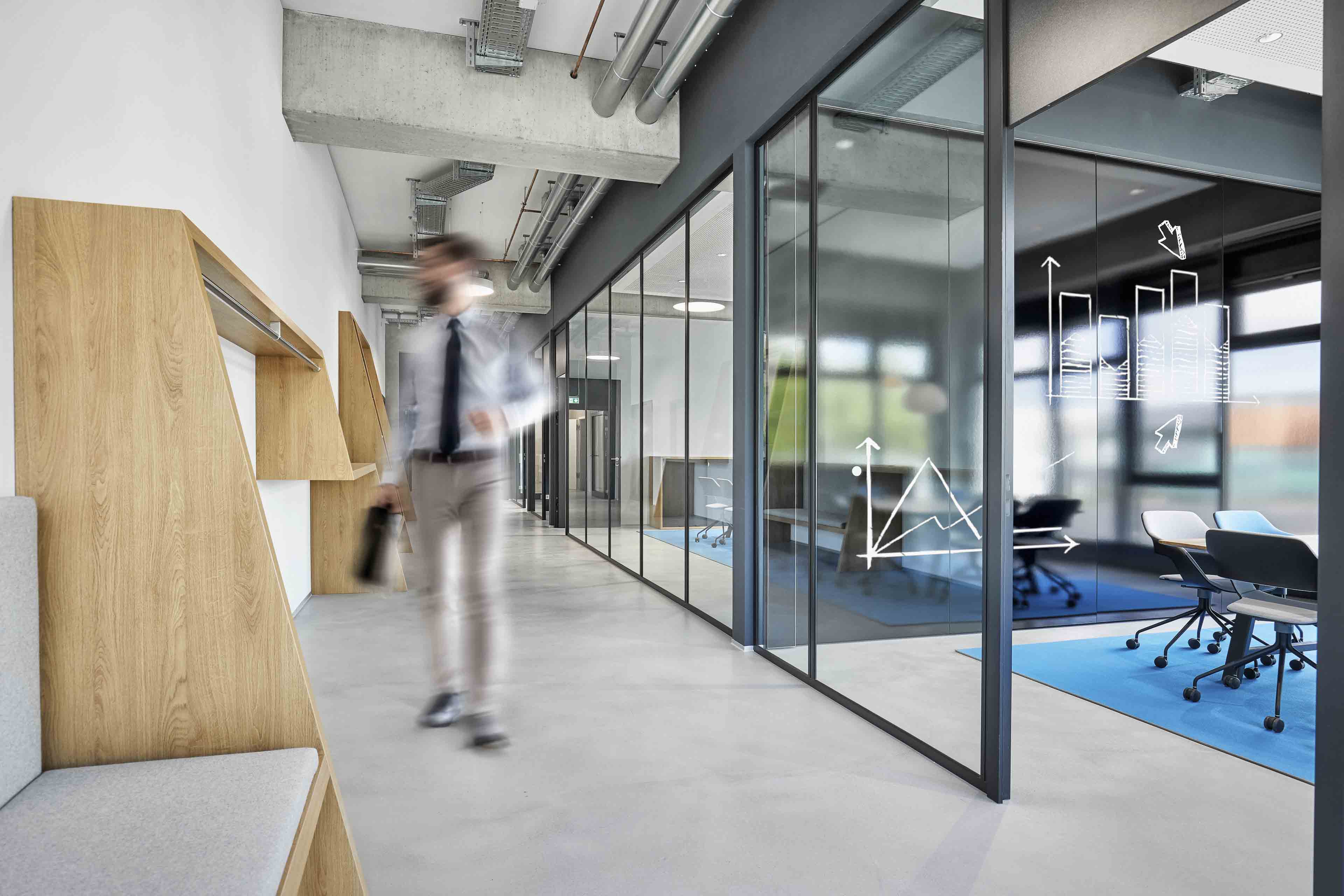 Geschäftsmann mit Aktentasche läuft durch modernen Büroflur mit Glaswänden und sichtbaren Diagrammen.