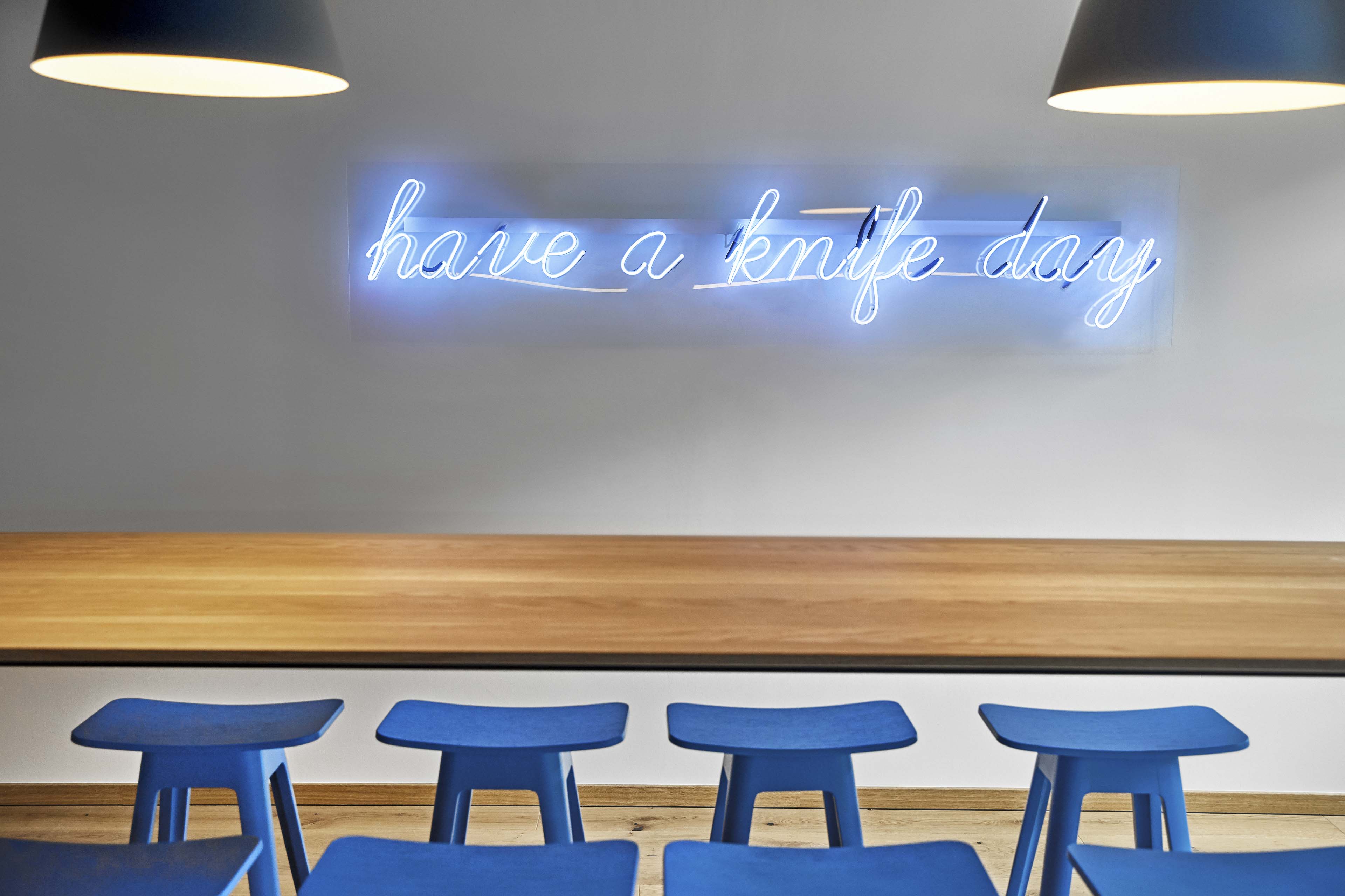 Neonlicht mit der Aufschrift 'have a knife day' über einer Holztheke und blauen Barhockern in einem modernen Raum.