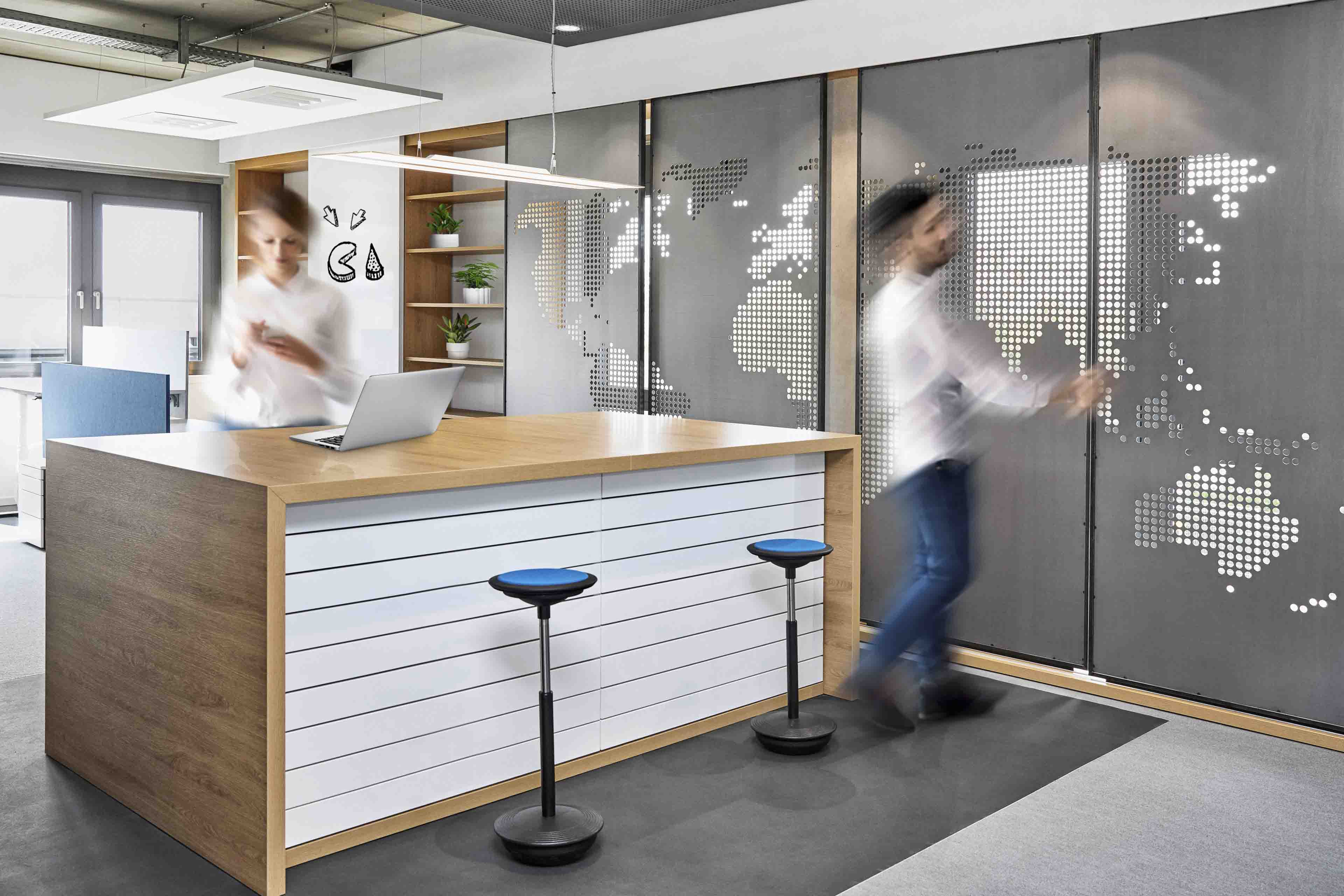 Modernes Büro mit zwei verschwommenen Personen, Stehtisch mit Laptop und zwei blauen Hockern vor einer Wand mit Weltkarten-Muster.