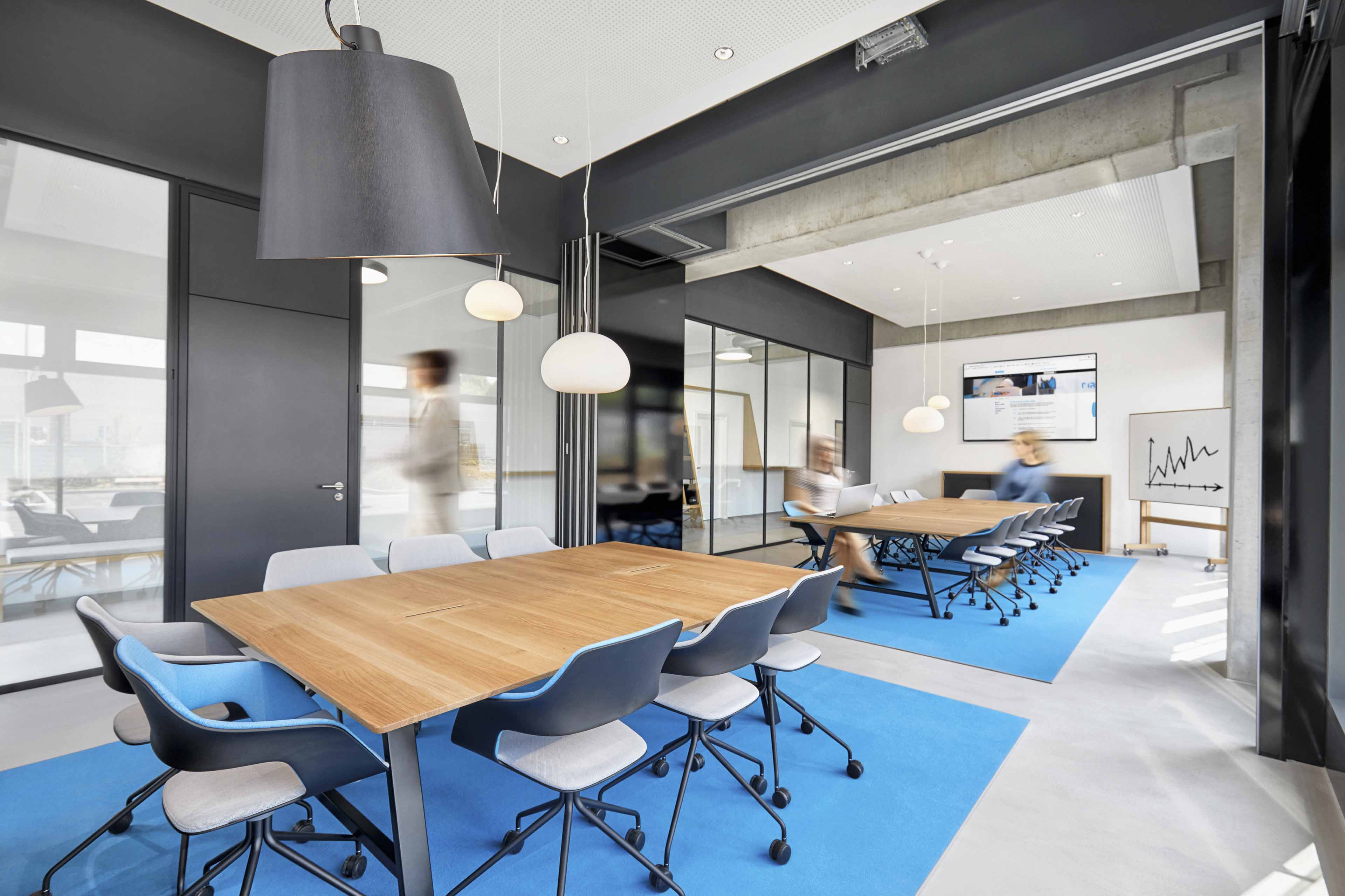 Modernes Besprechungszimmer mit zwei Holztischen auf blauen Teppichen, mehreren drehbaren Stühlen und drei verschwommenen Personen.