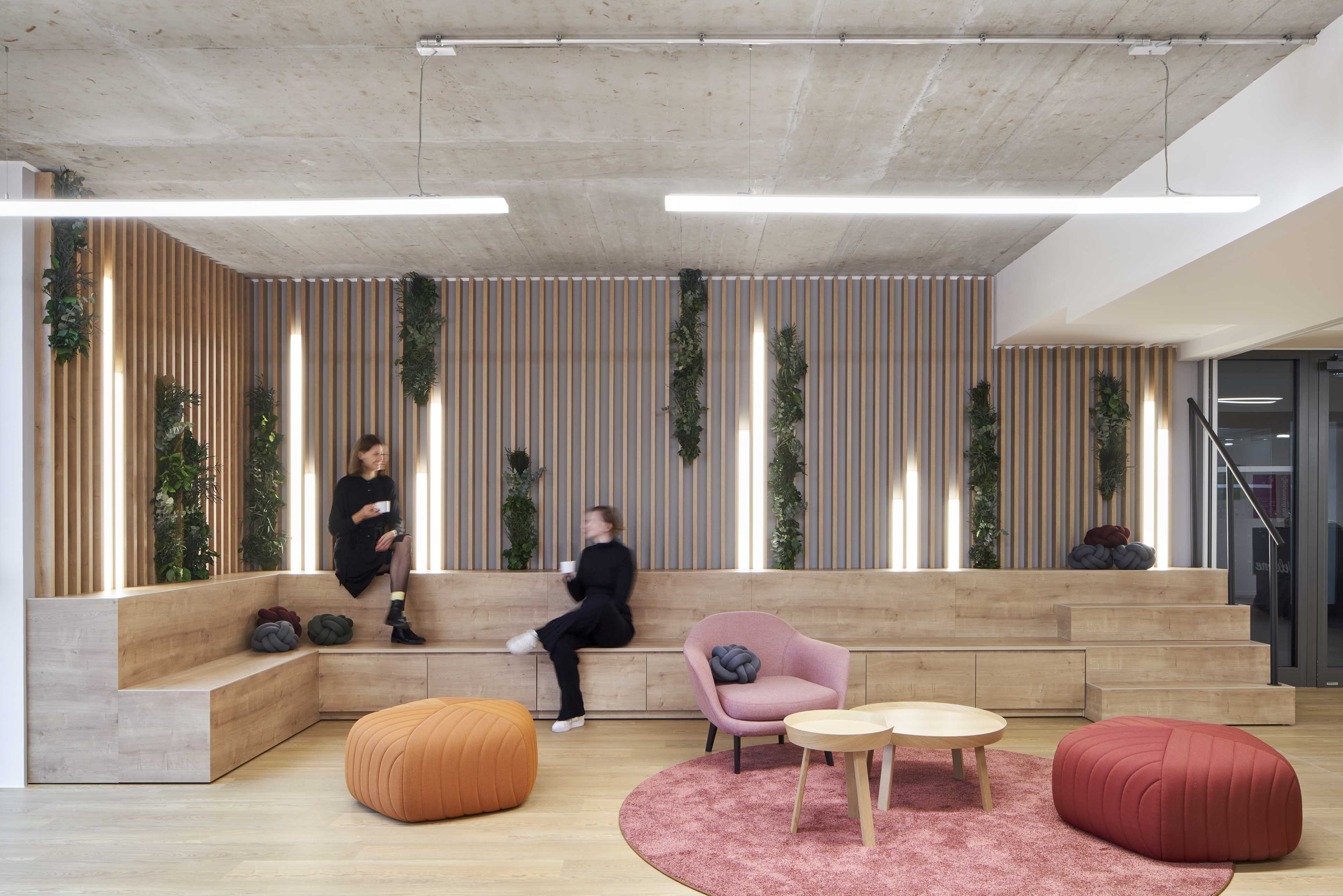 Moderner Lounge-Bereich mit hölzernen Sitzstufen, grünen Wandpflanzen, einem rosa Sessel, orangem und rotem Hocker sowie zwei niedrigen Holztischen auf einem rosa Teppich.