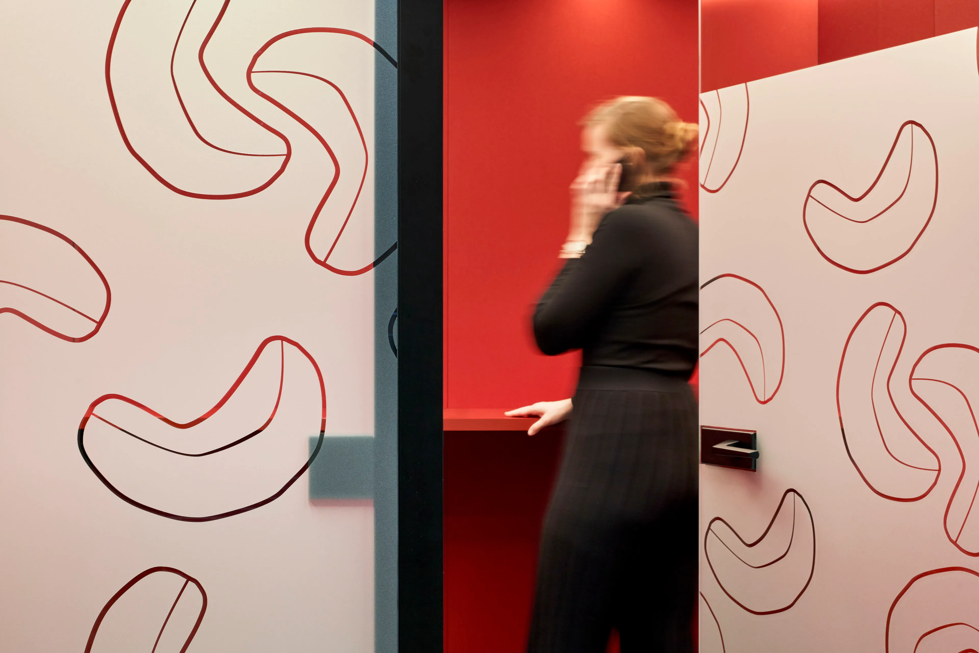 Verschwommene Frau im schwarzen Outfit spricht in farbenfroher Telefonkabine mit rot-weißen Wänden und Cashew-Nuss-Motiven.