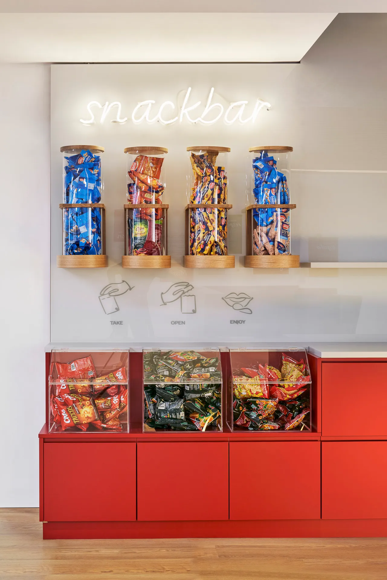Ein moderner Snackbar-Bereich mit vier hohen Glasbehältern voller Snacks an der Wand und drei Acrylbehältern mit verschiedenen Snacks auf einem roten Schrank.