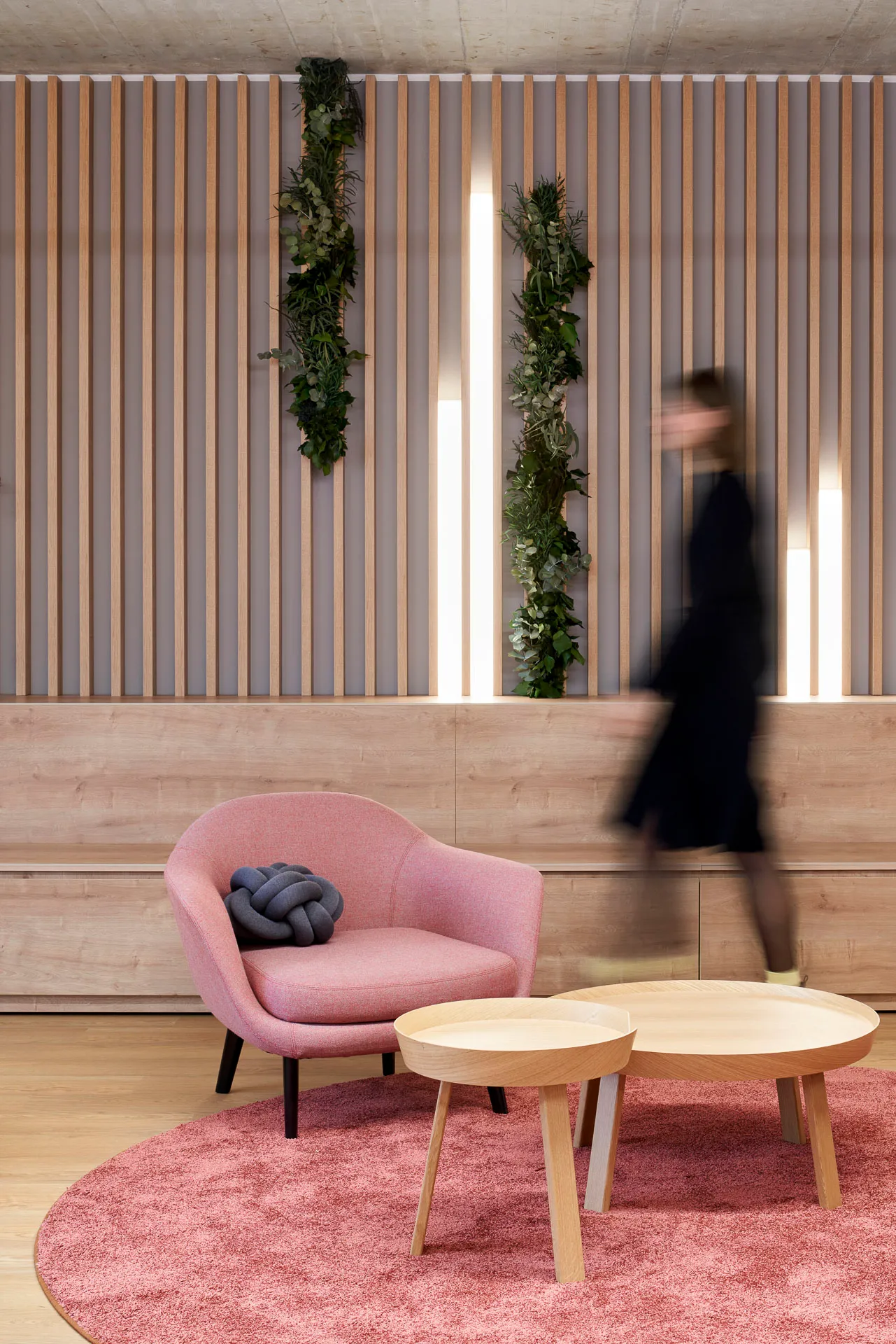Moderner Sitzbereich mit rosa Sessel, zwei runden Holztischen auf einem rosa Teppich, hängenden grünen Pflanzen an der Wand und einer verschwommenen Person, die vorbeigeht.