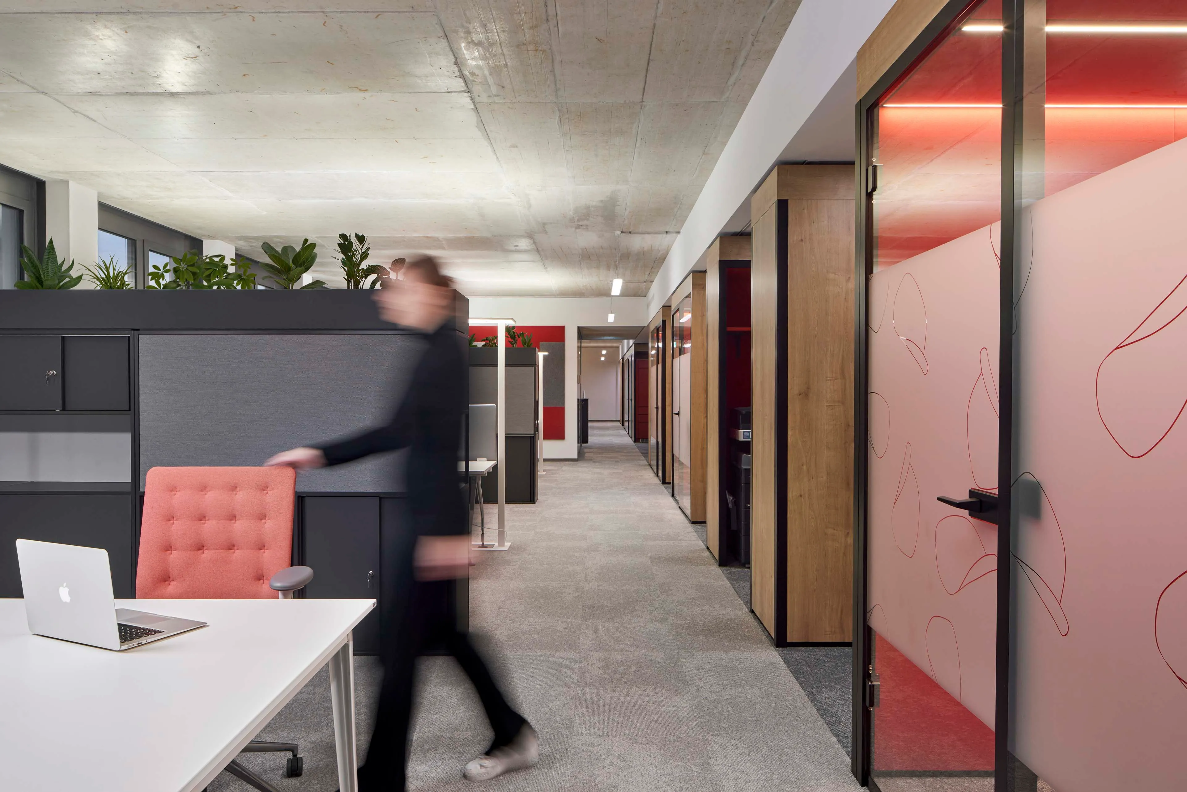 Moderne Büroumgebung mit Arbeitsplätzen auf der linken Seite, Glastüren mit roten Linienmustern rechts und einer Person in Bewegung.