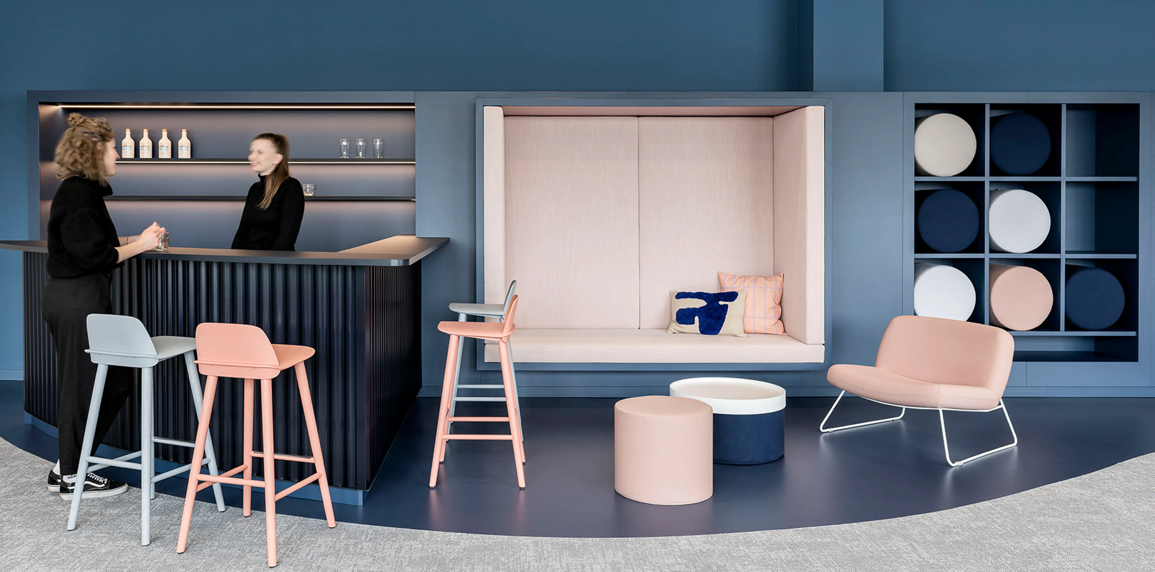 Moderne Lounge mit barblauem Tresen, rosa und grauen Barhockern, eingebauter Sitznische mit Kissen und gepolstertem Sofa sowie Wandregal mit runden Kissen.