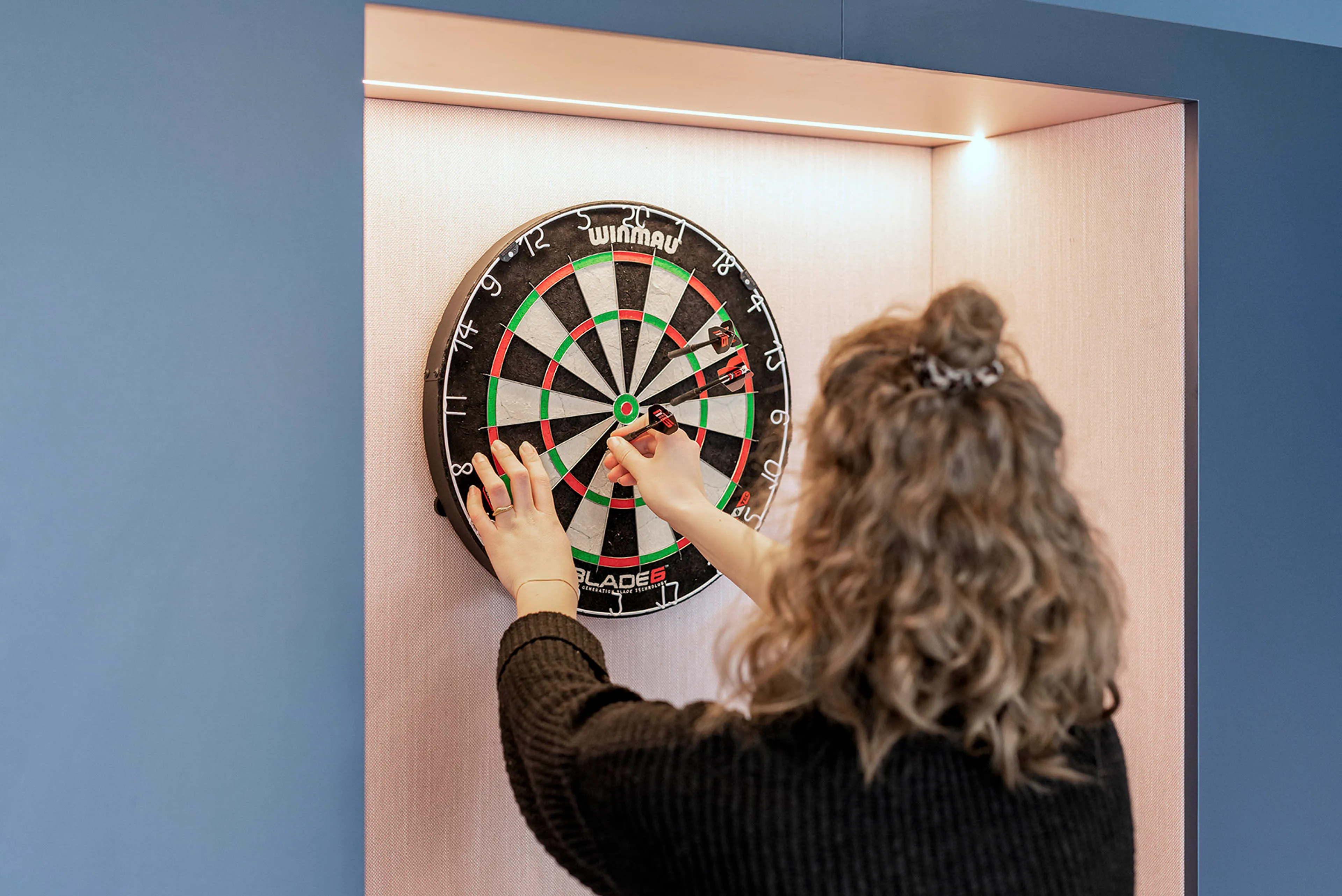 Eine Frau steckt Darts auf ein beleuchtetes Dartboard in einer Wandnische.
