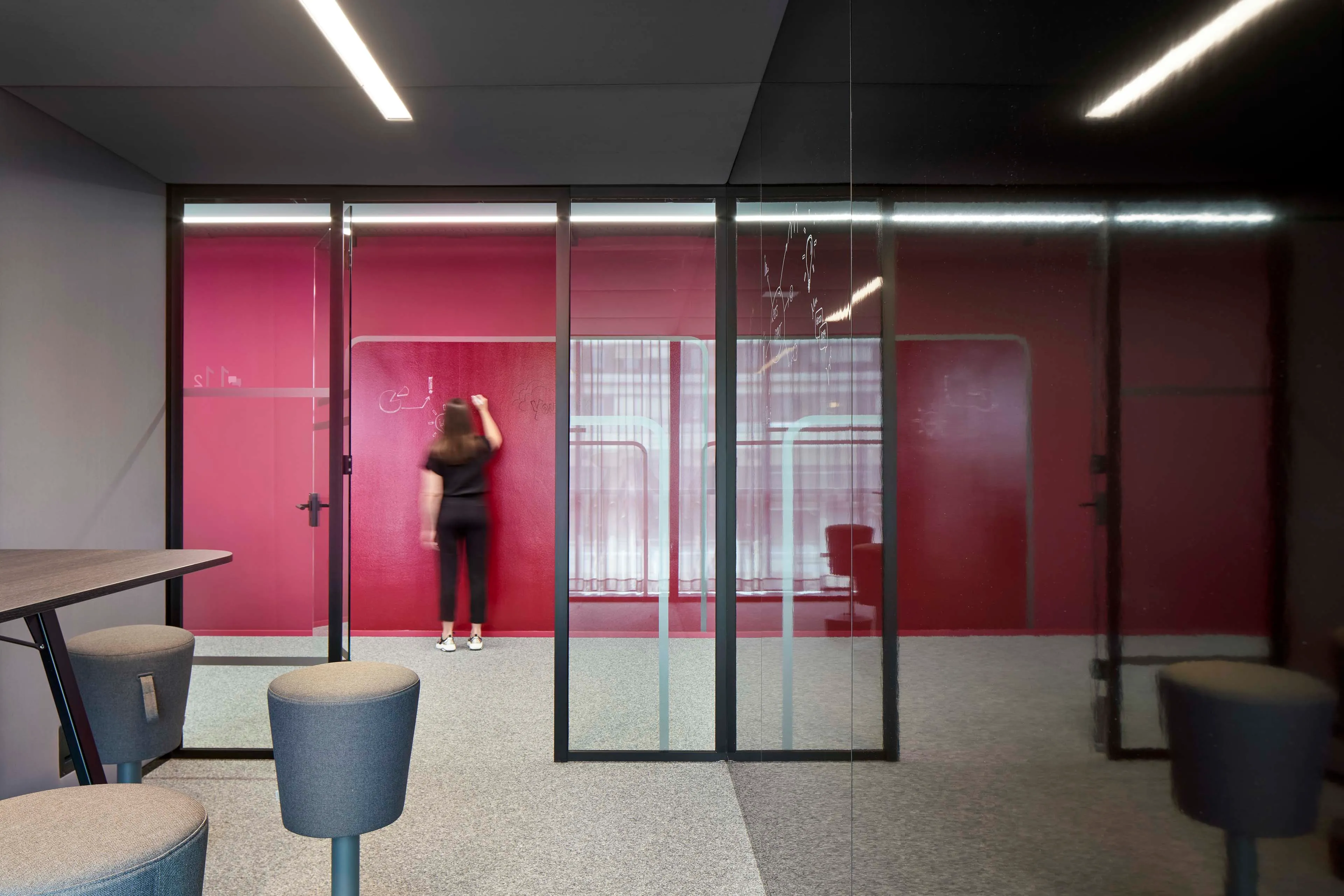 Frau, die an einer roten Wand in einem modernen Büro schreibt, umgeben von Glaswänden und grauen Sitzhockern.
