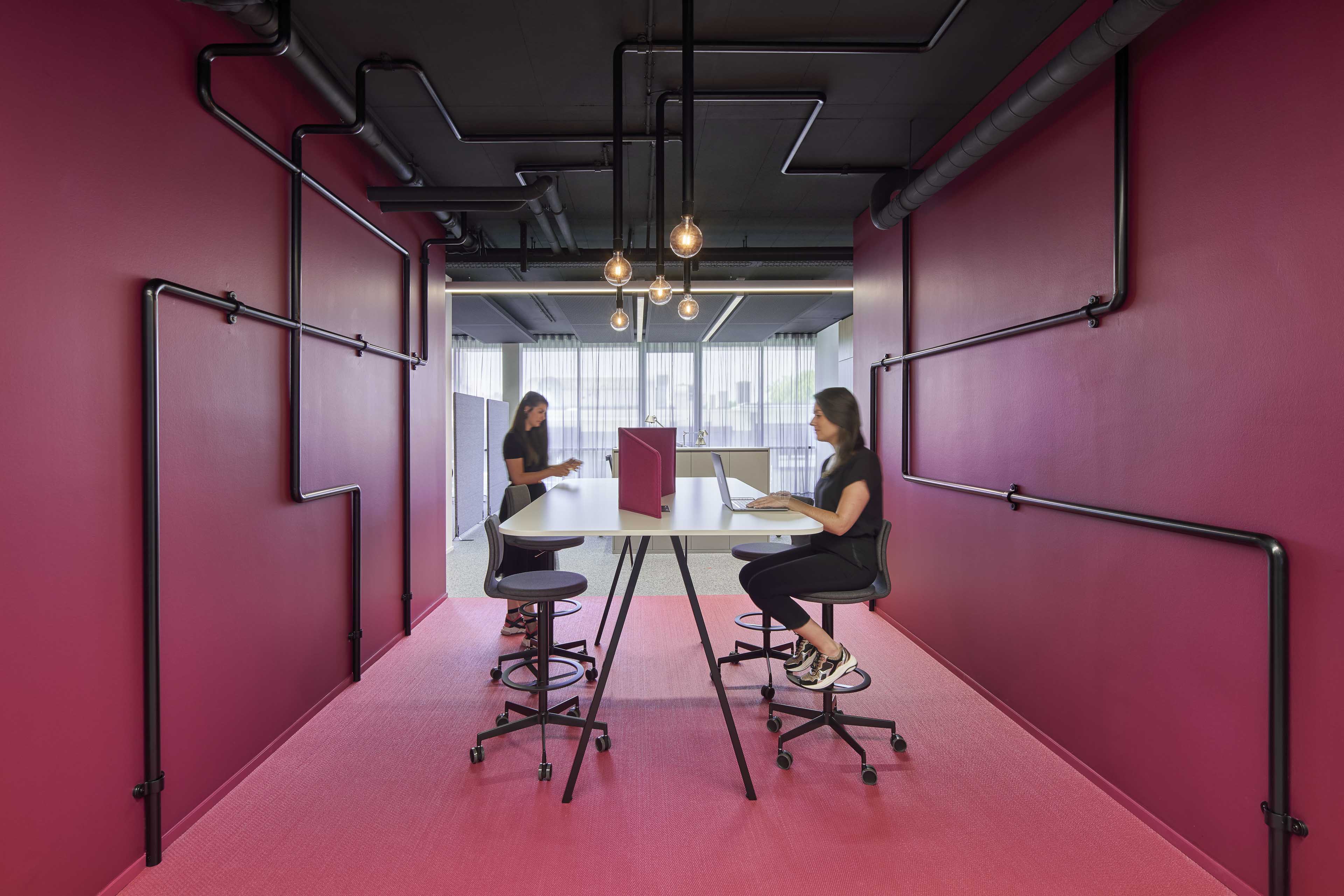 Zwei Frauen arbeiten an einem langen Tisch mit Trennwänden in einem modernen Büro mit rosa Wänden und Teppich, sichtbare schwarze Rohre an den Wänden.