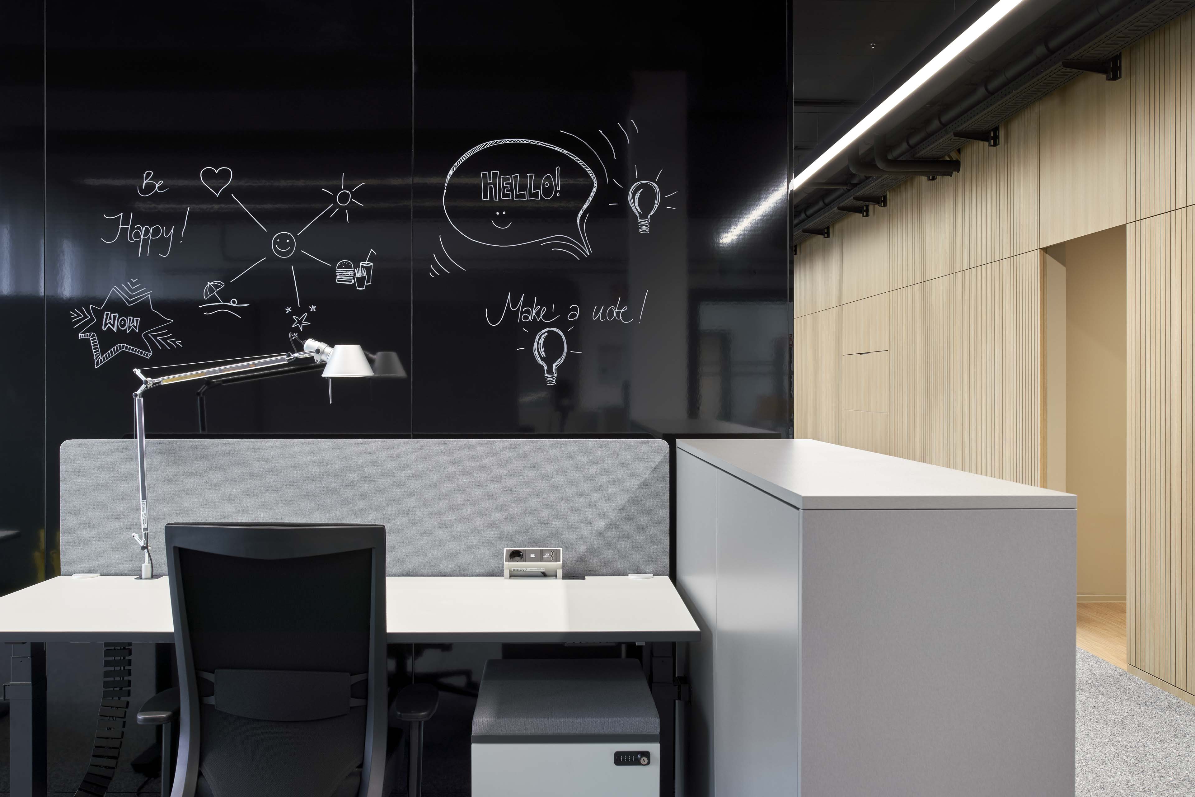 Moderner Arbeitsplatz mit Schreibtisch, Schreibtischlampe und Stuhl vor einer schwarzen Glaswand mit weißen Zeichnungen und motivierenden Worten.