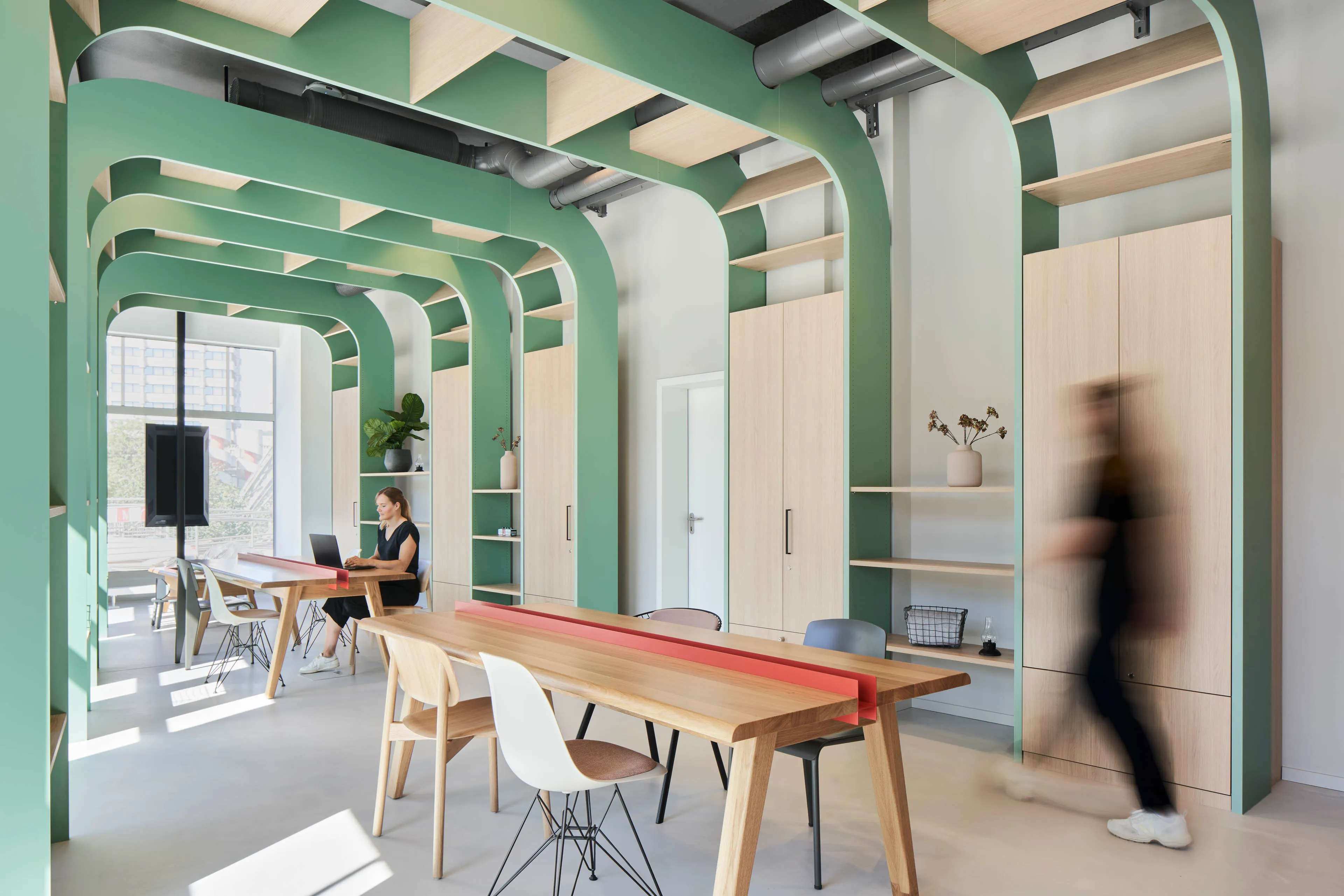 Modernes Büro mit grünen Stahlträgern, Holzregalen und -schränken, mehreren Holz- und Kunststoffstühlen und zwei Personen, von denen eine am Laptop arbeitet und die andere vorbeigeht.