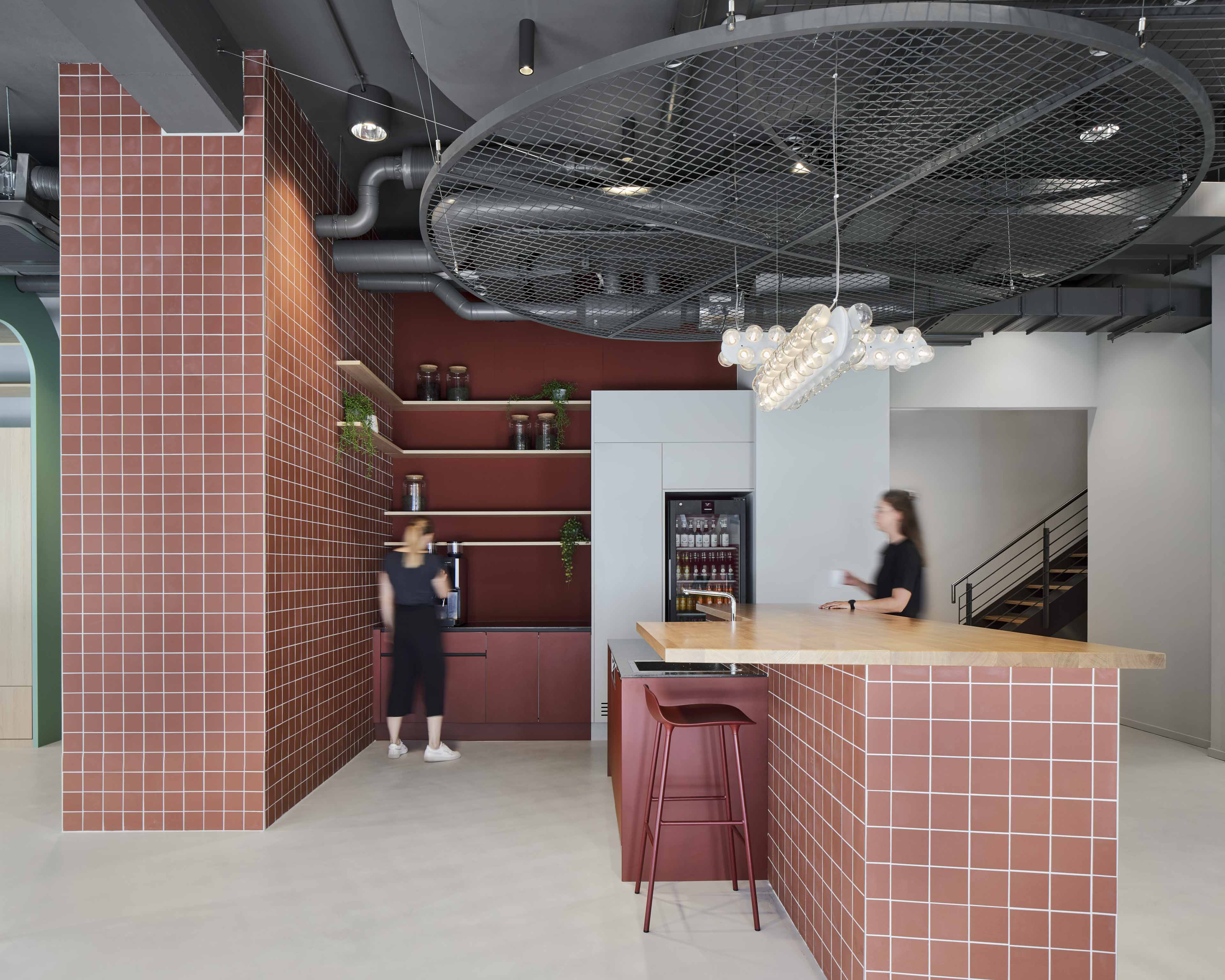 Moderne Büro-Küche mit terrakottafarbenen Kacheln, Holztresen, zwei verschwommenen Personen und einer großen runden Deckenbeleuchtung.
