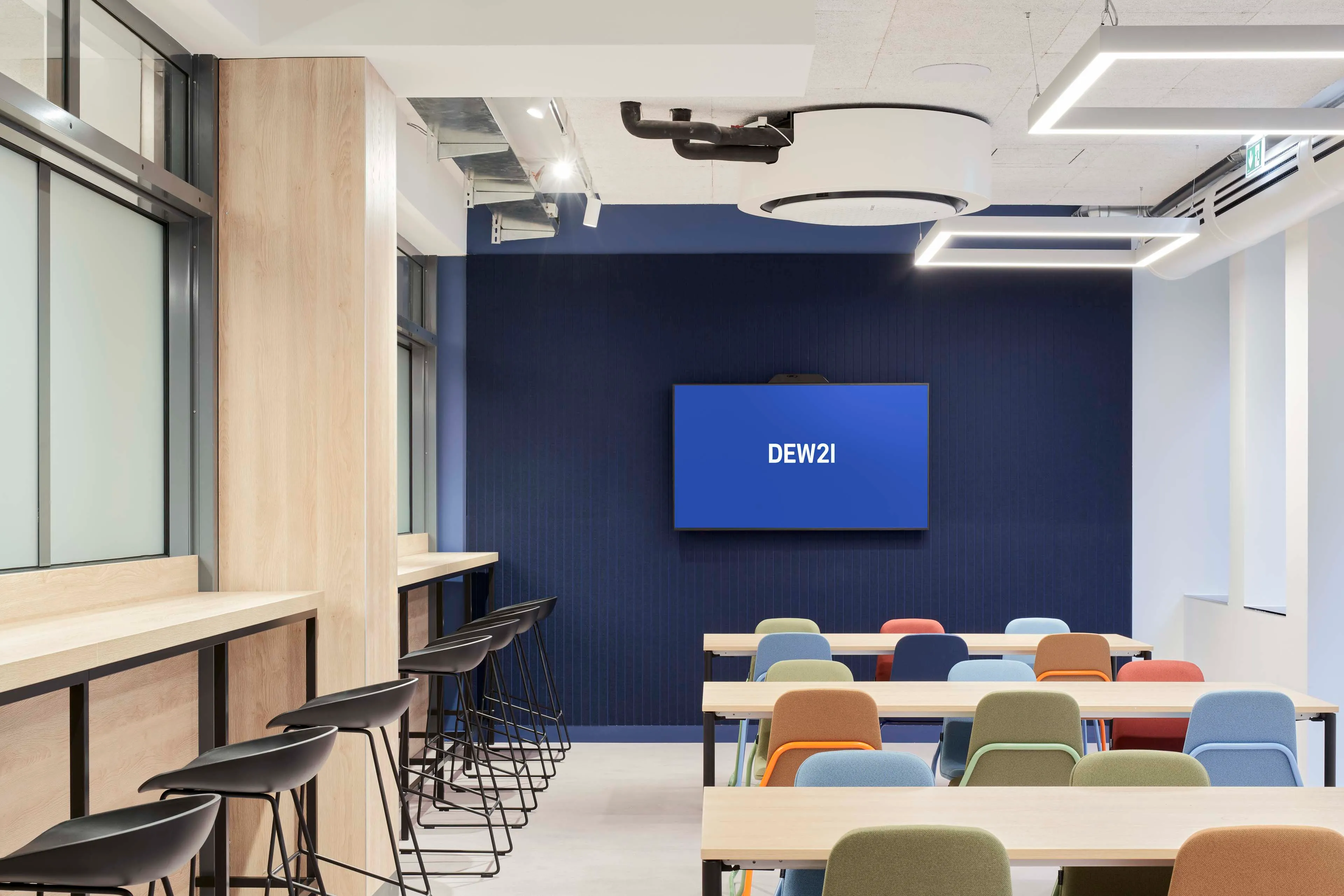 Moderner Konferenzraum mit bunten Stühlen, Holz Tischen und einem Bildschirm mit der Aufschrift DEW21 an der dunkelblauen Wand.