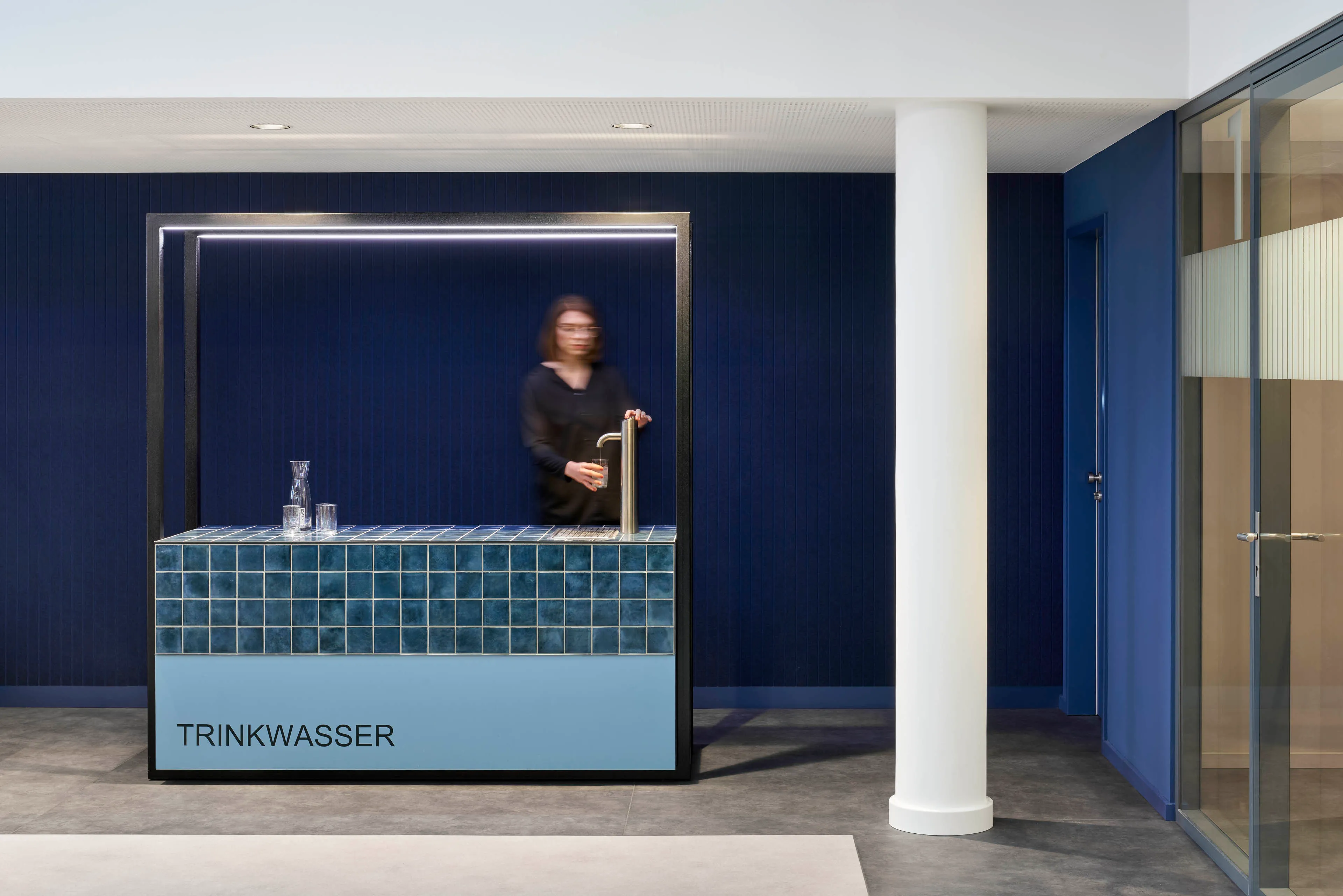 Moderner Trinkwasserbrunnen mit blauen Fliesen in einem Raum mit dunkelblauer Wand und weißer Säule, Person mit Glas am Wasserhahn.