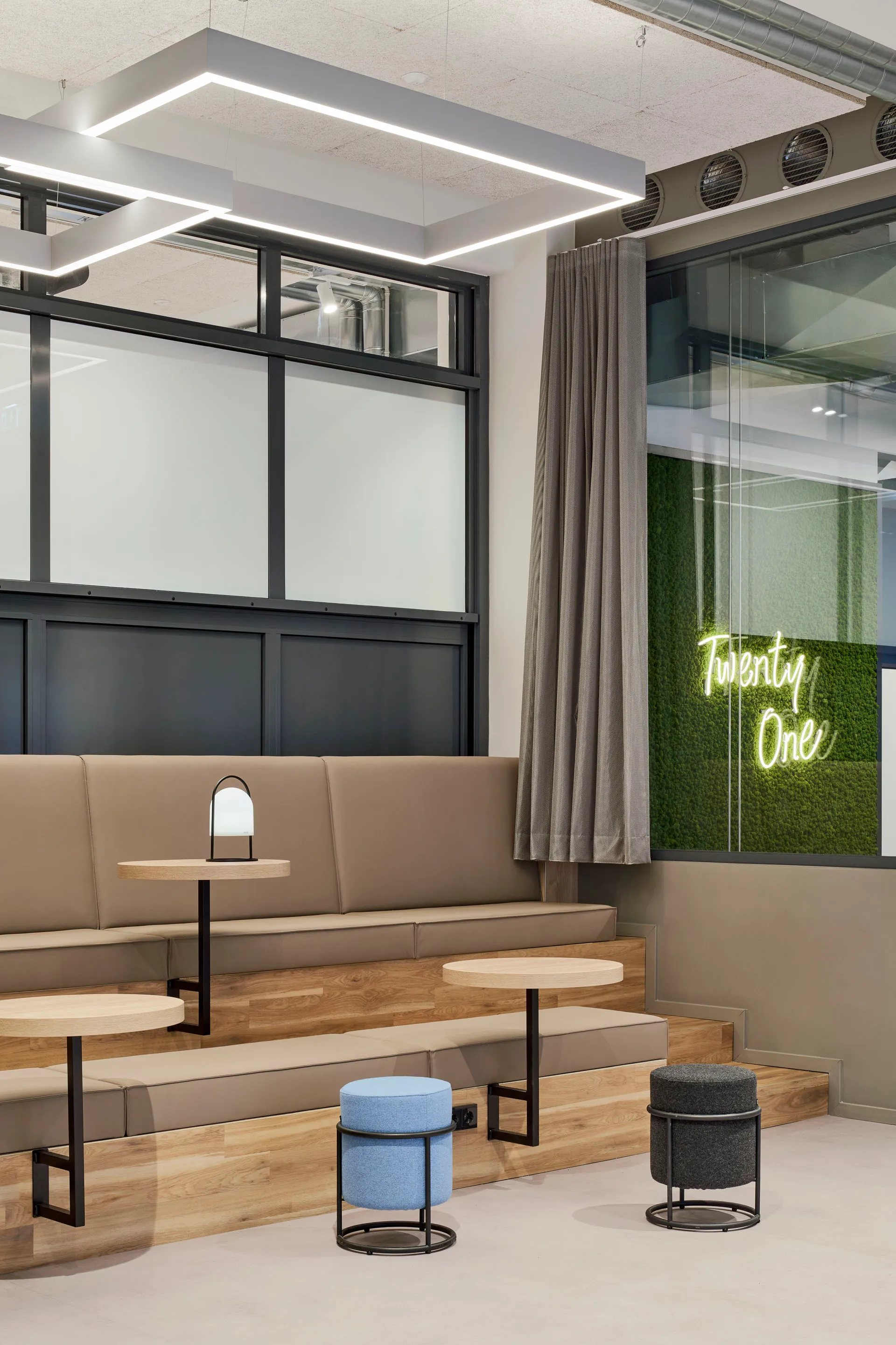 Moderner Sitzbereich mit zweistufigen beigen Polstersitzen, kleinen runden Holztischen und zwei gepolsterten Hockern, neonbeleuchteter Schriftzug "Twenty One" an der Wand.