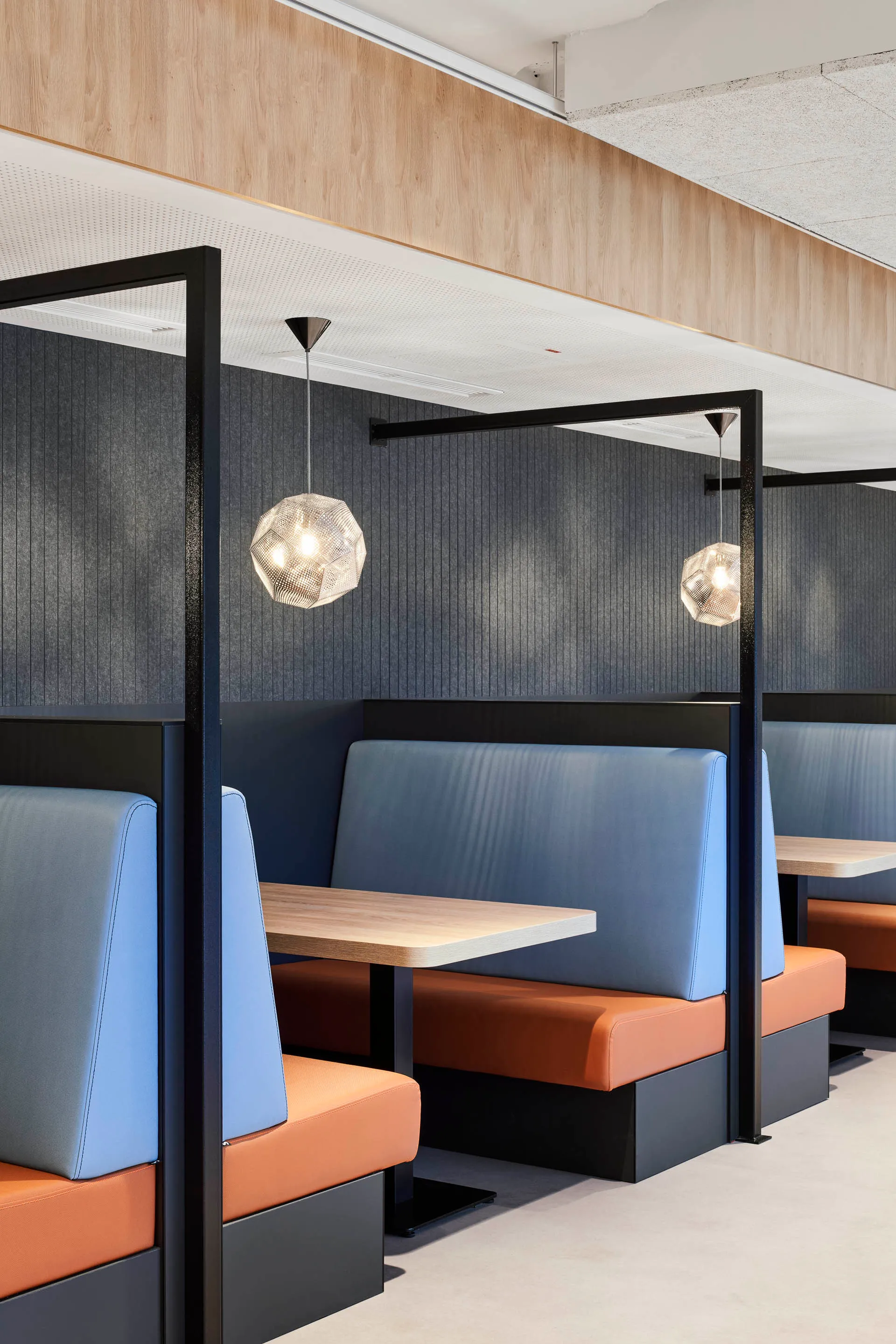 Moderne Restaurant-Nischen mit blauen Rückenlehnen, orangefarbenen Sitzpolstern und hölzernen Tischen unter geometrischen Pendelleuchten.