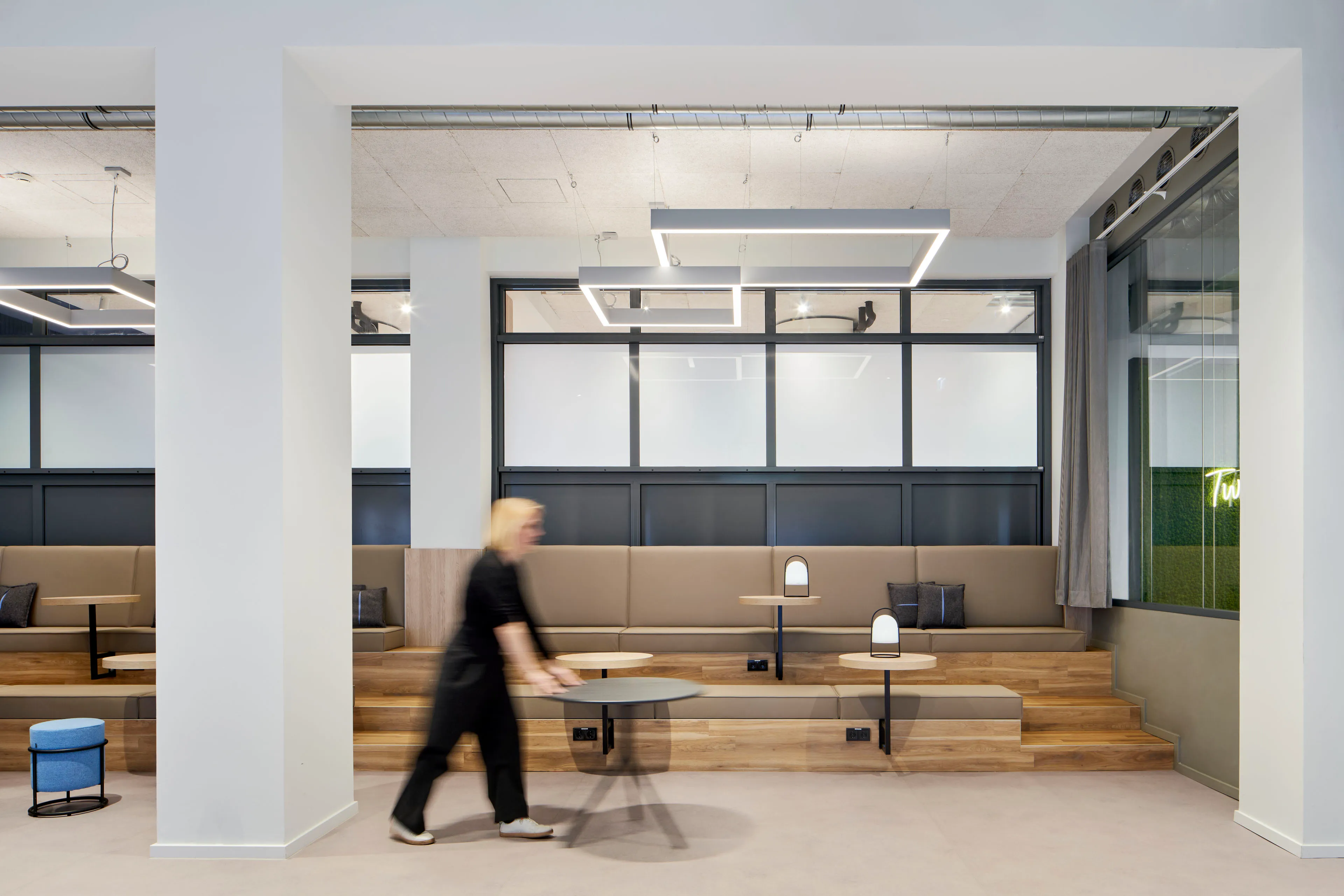 Moderne Sitzterrasse mit Holzstufen, beigem Polstersofa, kleinen Tischen und abstrahierten Deckenleuchten in einem hellen Raum.