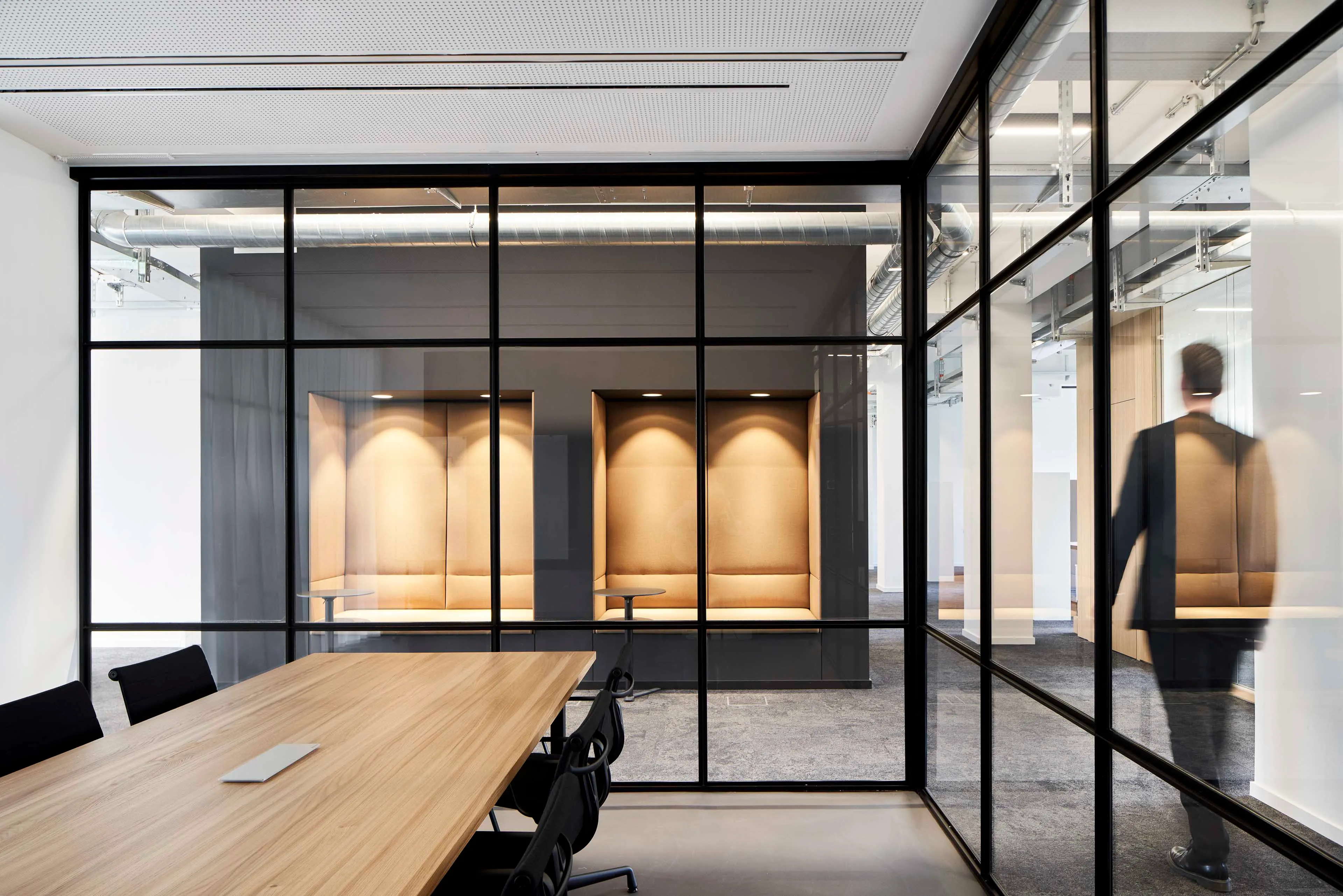 Modernes Büro mit einem Besprechungstisch, komfortablen Sitznischen hinter Glaswänden und einem Mann im Anzug, der vorbeigeht.