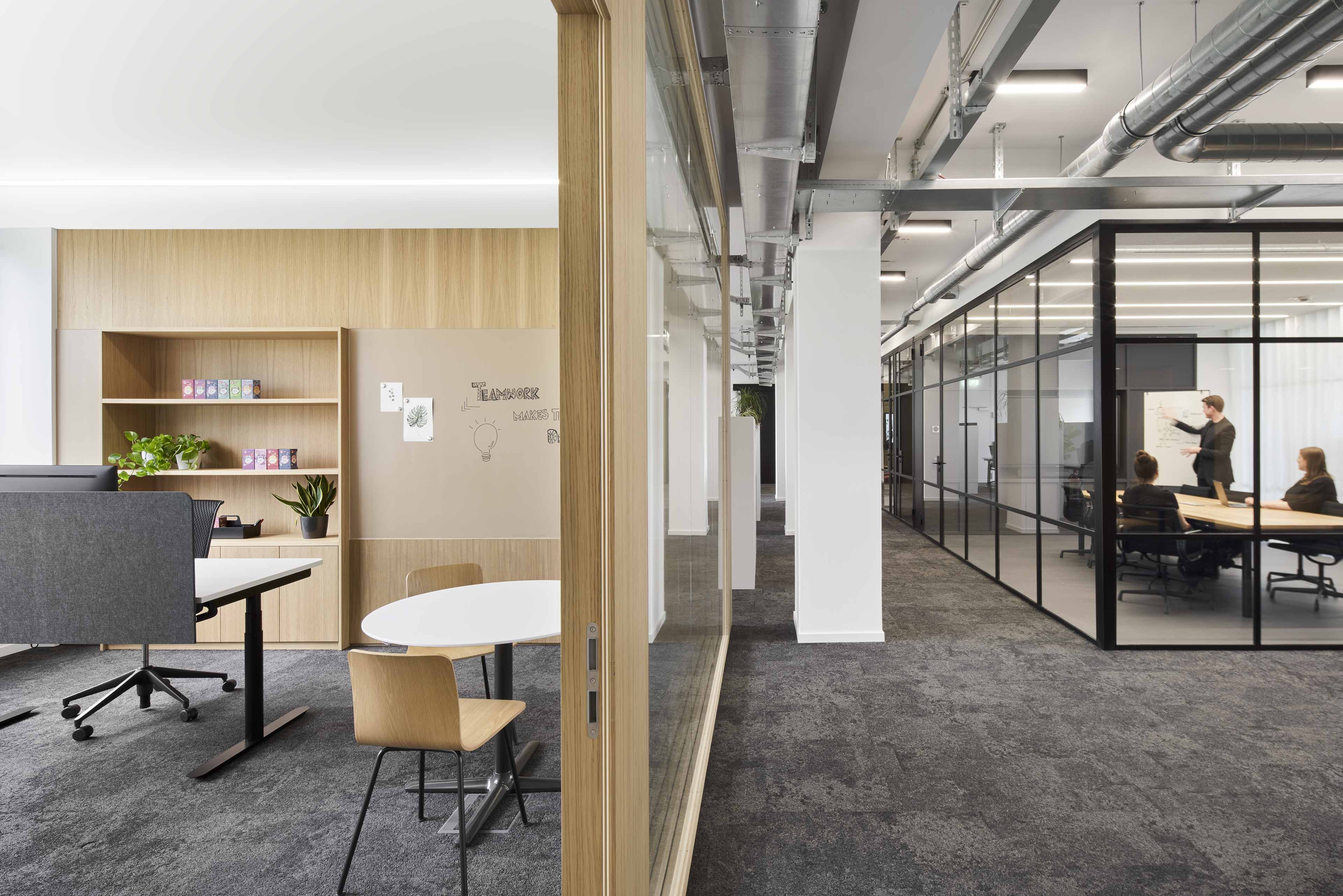 Modernes Büro mit Holzregalen, Pflanzen, Besprechungsraum mit Glaswänden und Personen bei einer Präsentation.