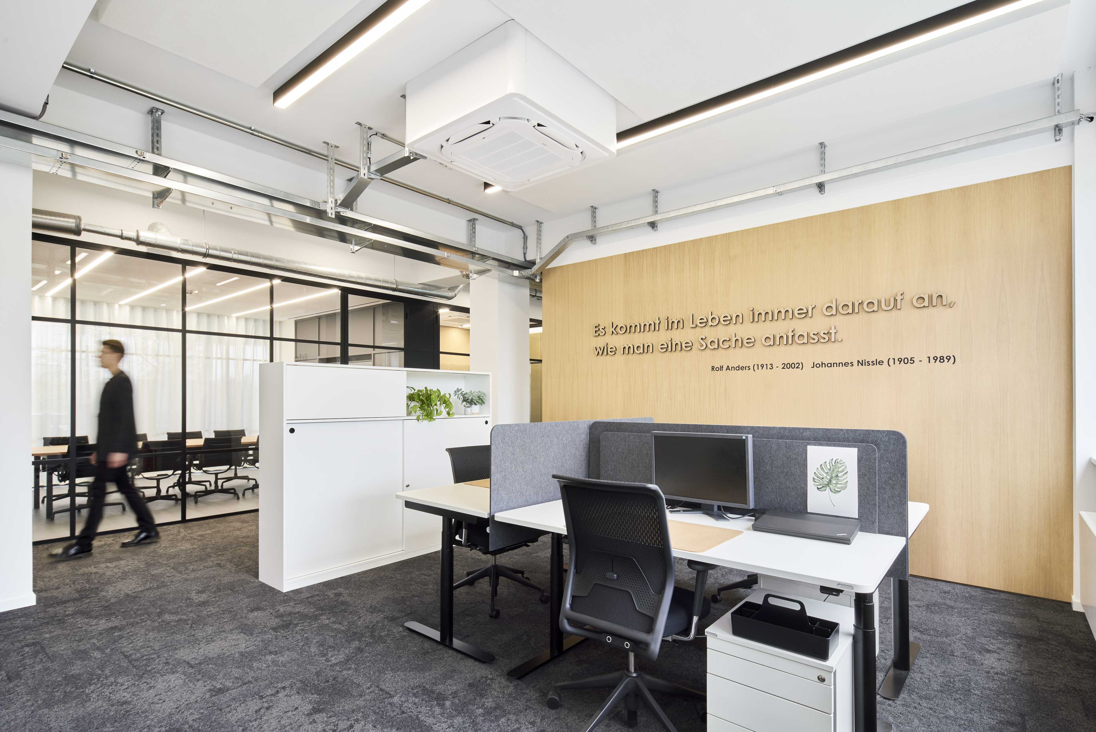 Moderner Büroraum mit Schreibtischen, Computern, Sichtschutzwänden und einem Mann, der an einer Glastrennwand zum Konferenzraum vorbeigeht.