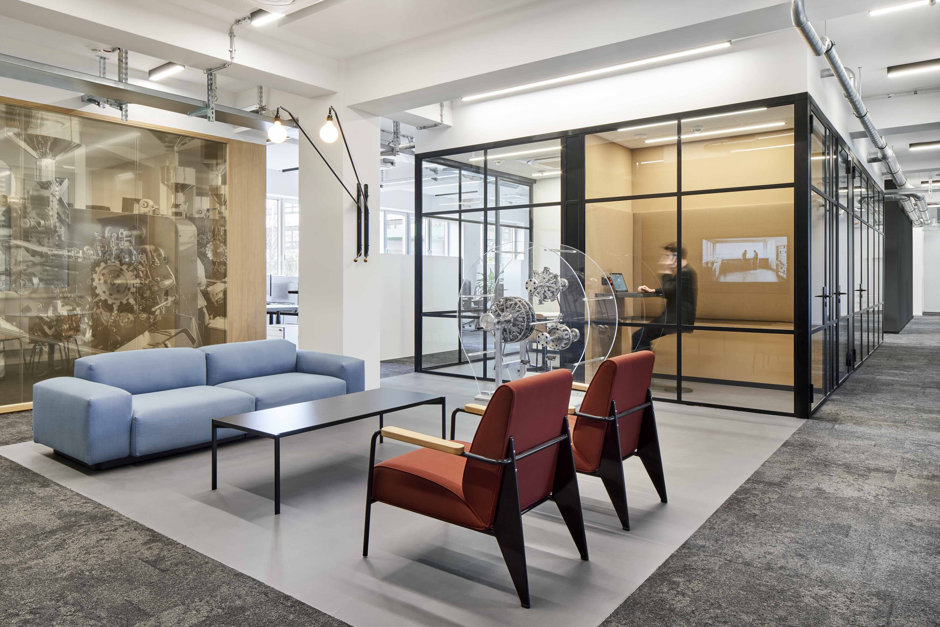 Modernes Büro mit blauem Sofa, zwei roten Sesseln, schwarzem Tisch und einem Besprechungsraum mit Glaswänden.