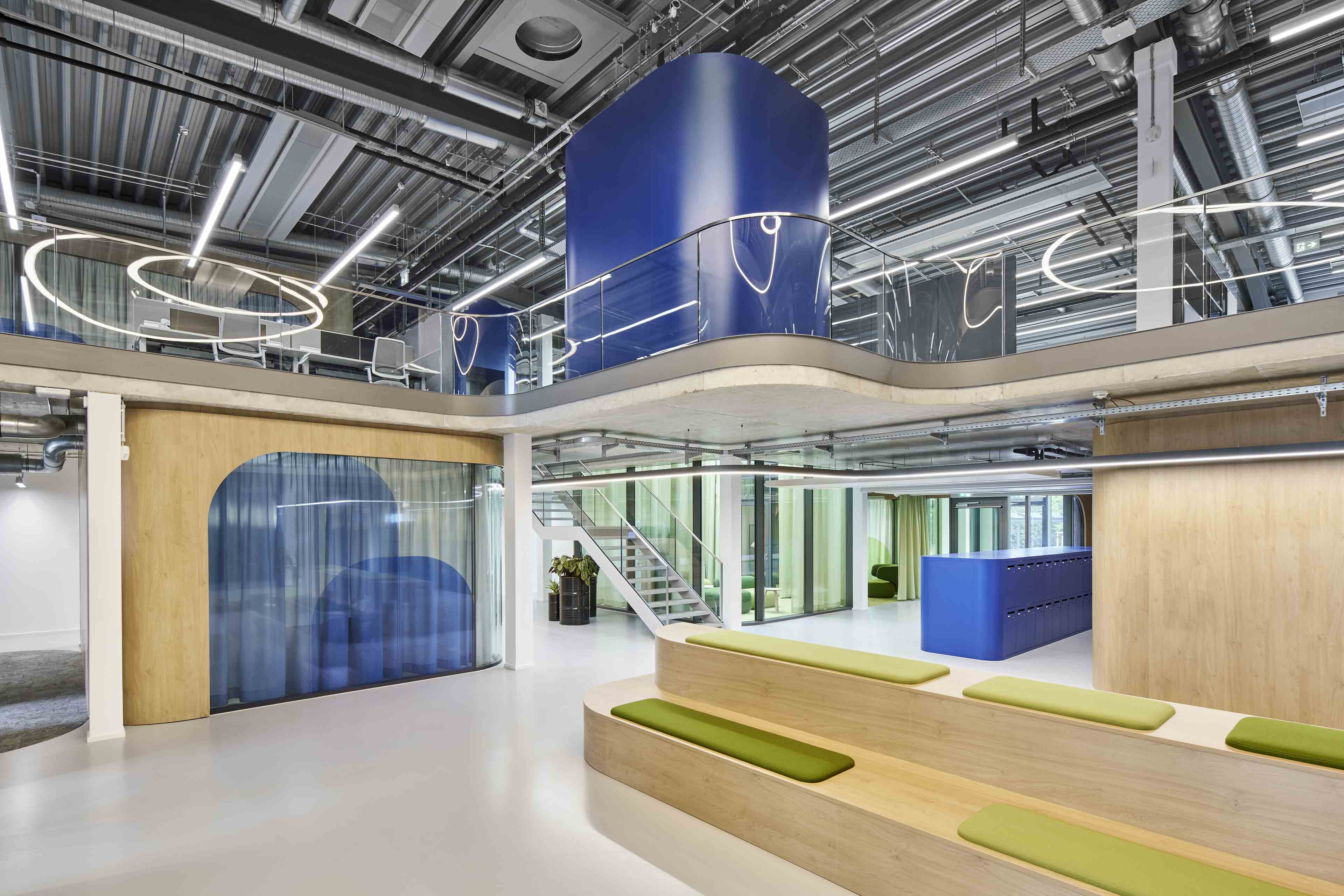 Modernes, helles Büro mit offener, zweistöckiger Raumarchitektur, blauen und grünen Akzenten und Sitzstufen aus Holz mit grünen Polstern.