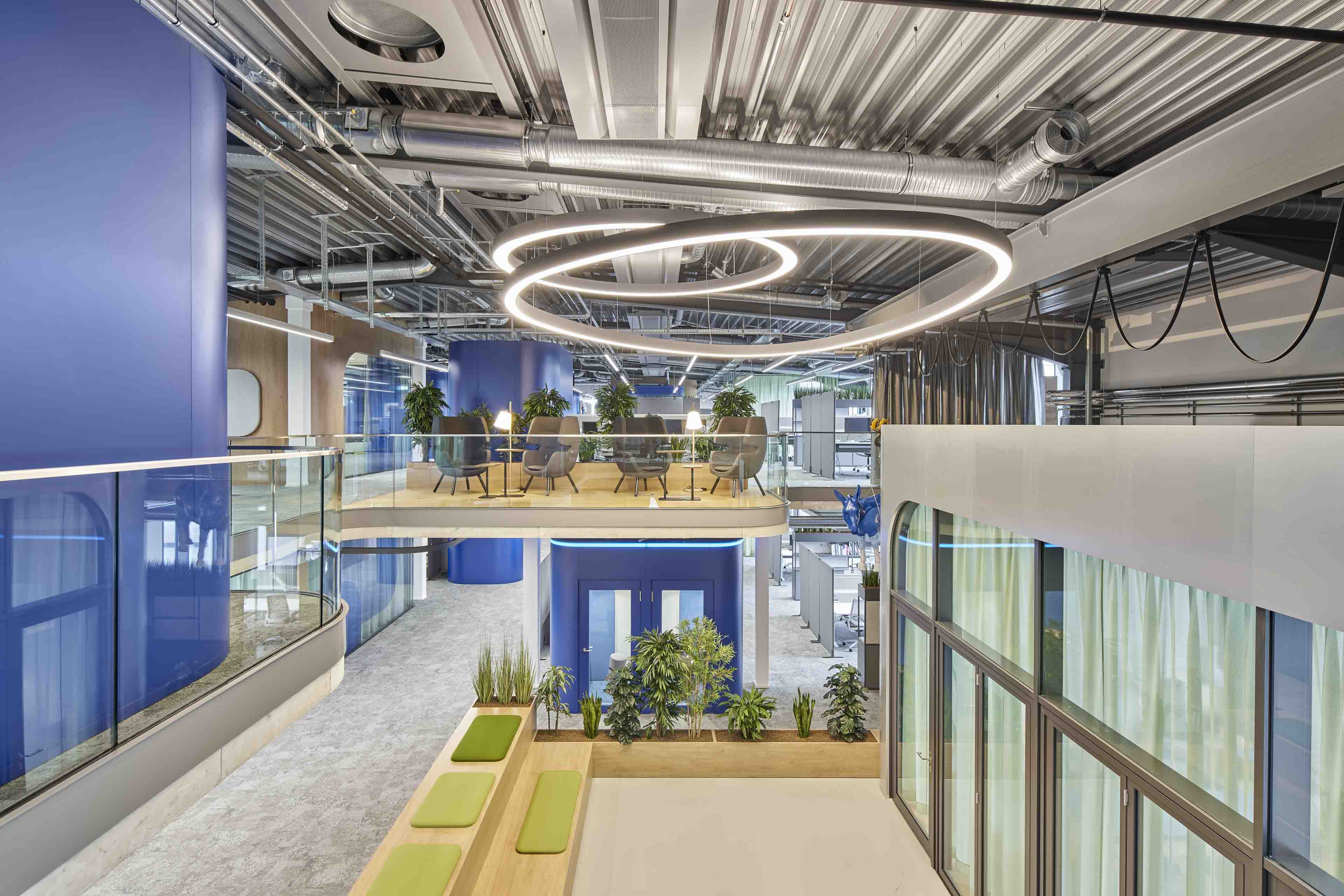Modernes Büro mit offenen Arbeitsbereichen, hängenden kreisförmigen Lichtinstallationen, blau lackierten Säulen und Pflanzen.